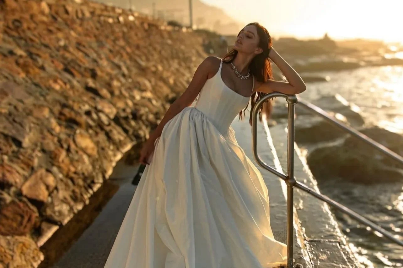 A woman in a white dress standing by the seaside at sunset, leaning on a metal railing.