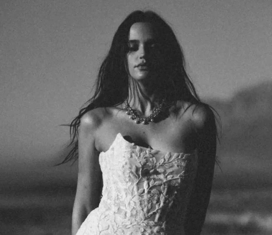 A black and white photo of a woman with long dark hair, wearing a strapless, textured dress and a necklace, standing outdoors during dusk or night with the sky in the background.