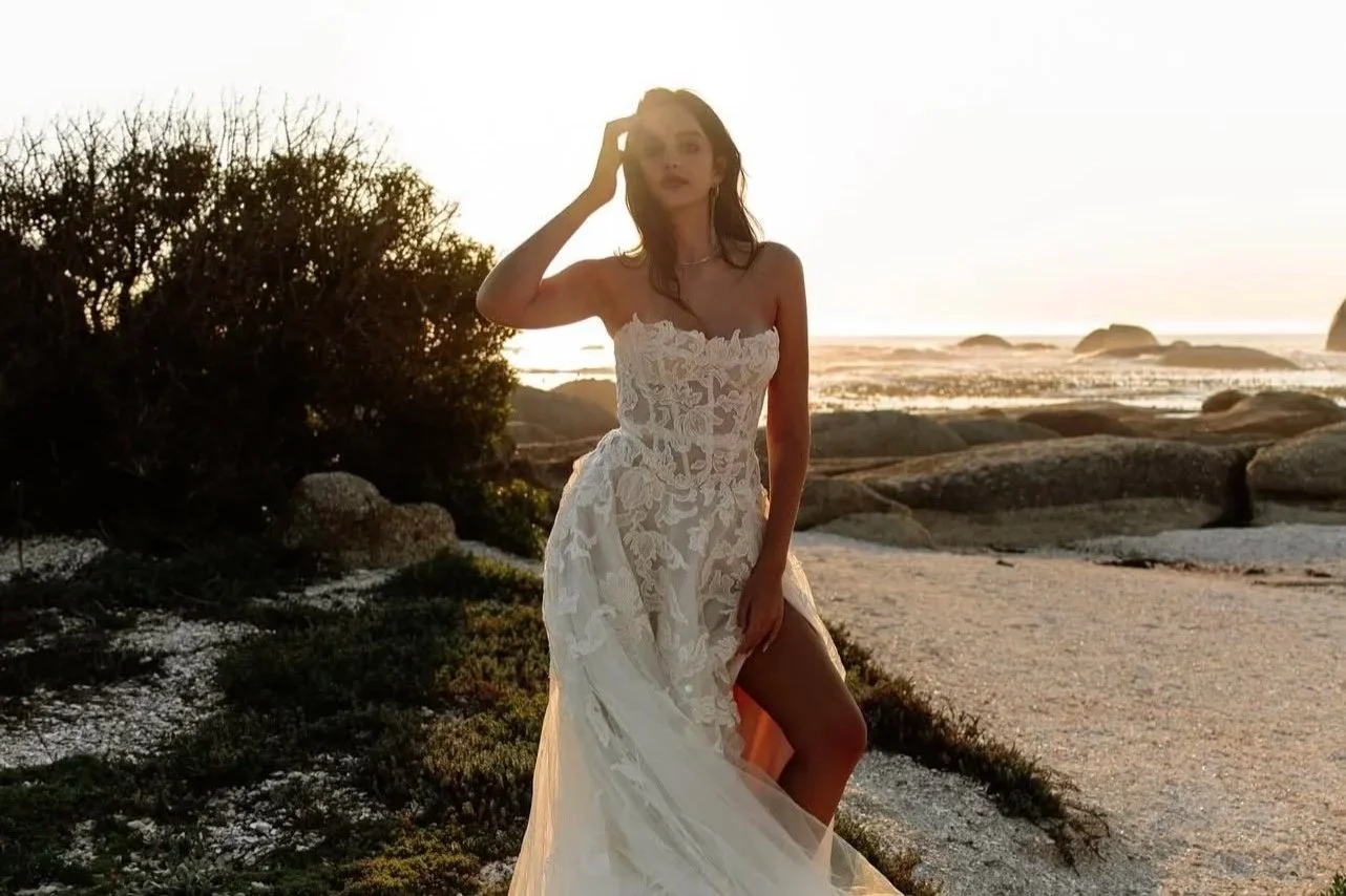 A woman in a white lace dress standing on a beach at sunset near rocks and bushes.