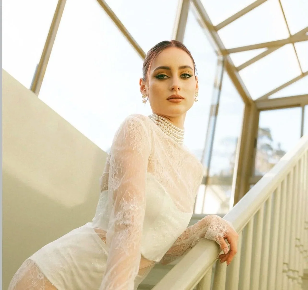 A woman in a lace dress and pearl jewelry posing on a staircase with glass windows in the background.