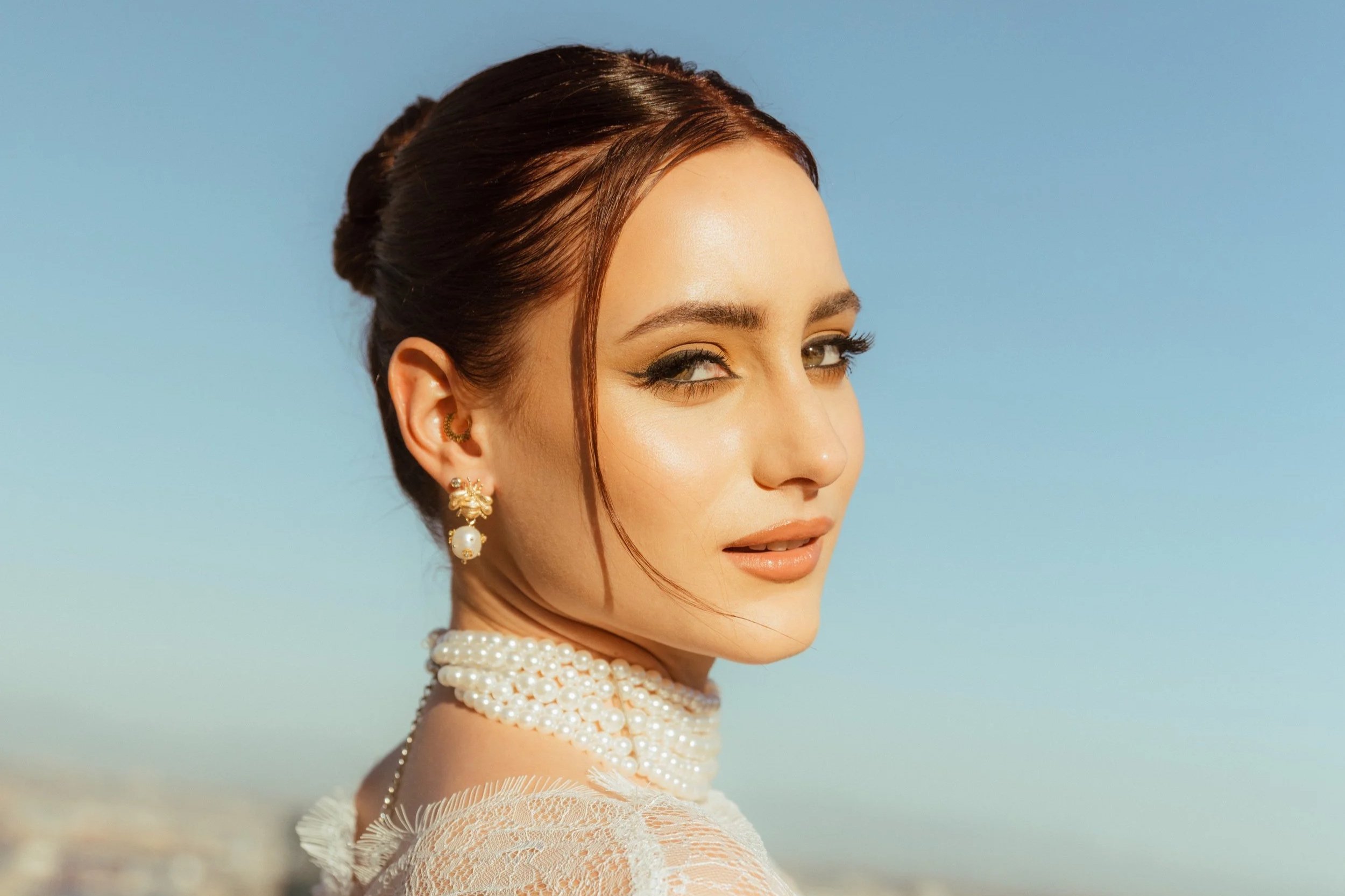 A woman with brown hair styled in an elegant updo, wearing pearl jewelry and a lace top, standing outdoors against a clear blue sky.