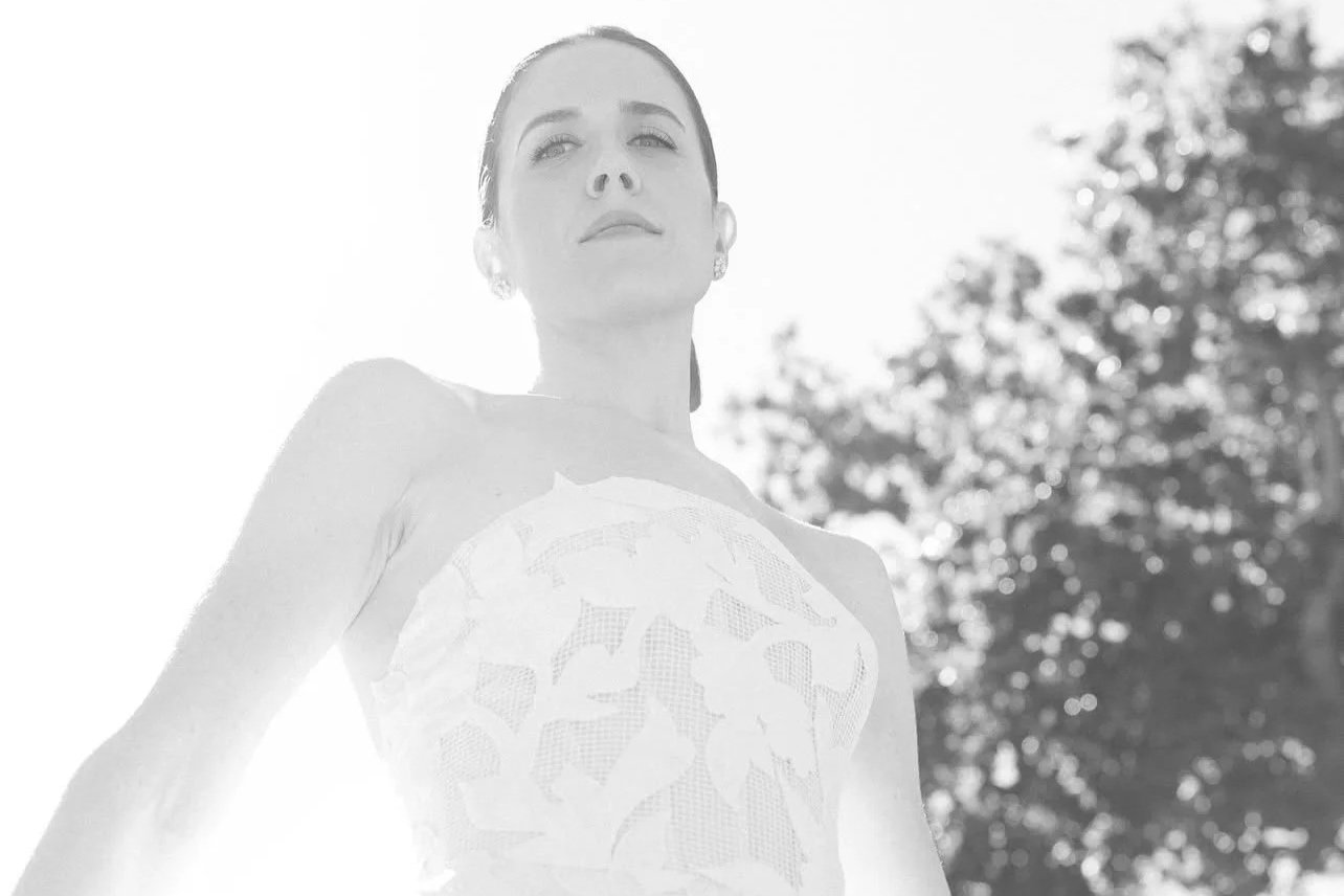 Black and white photo of a woman looking upward with sunlight behind her, standing outdoors with trees in the background.