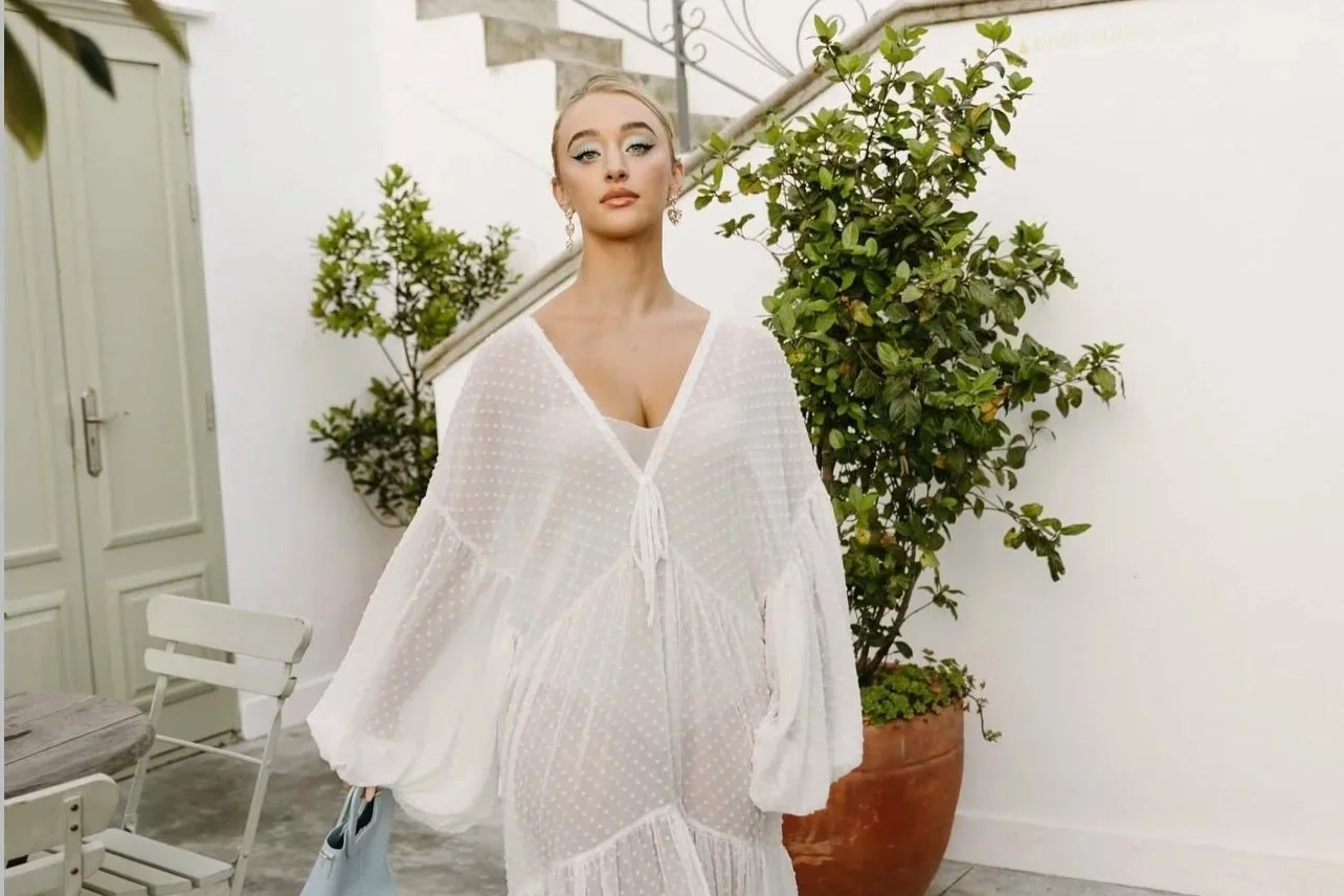 A woman in Cape Town in  a sheer white dress with a deep V neckline stands outdoors in front of greenery and potted plants.