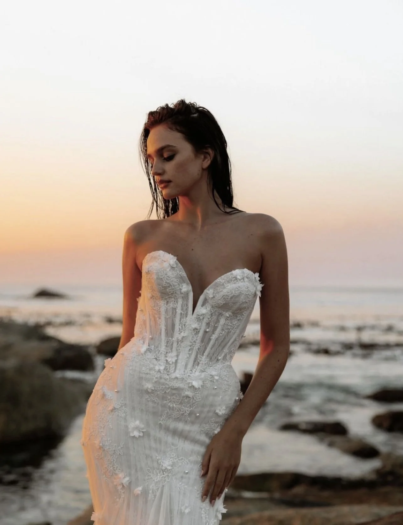 A woman in a white wedding dress standing on a rocky beach during sunset.