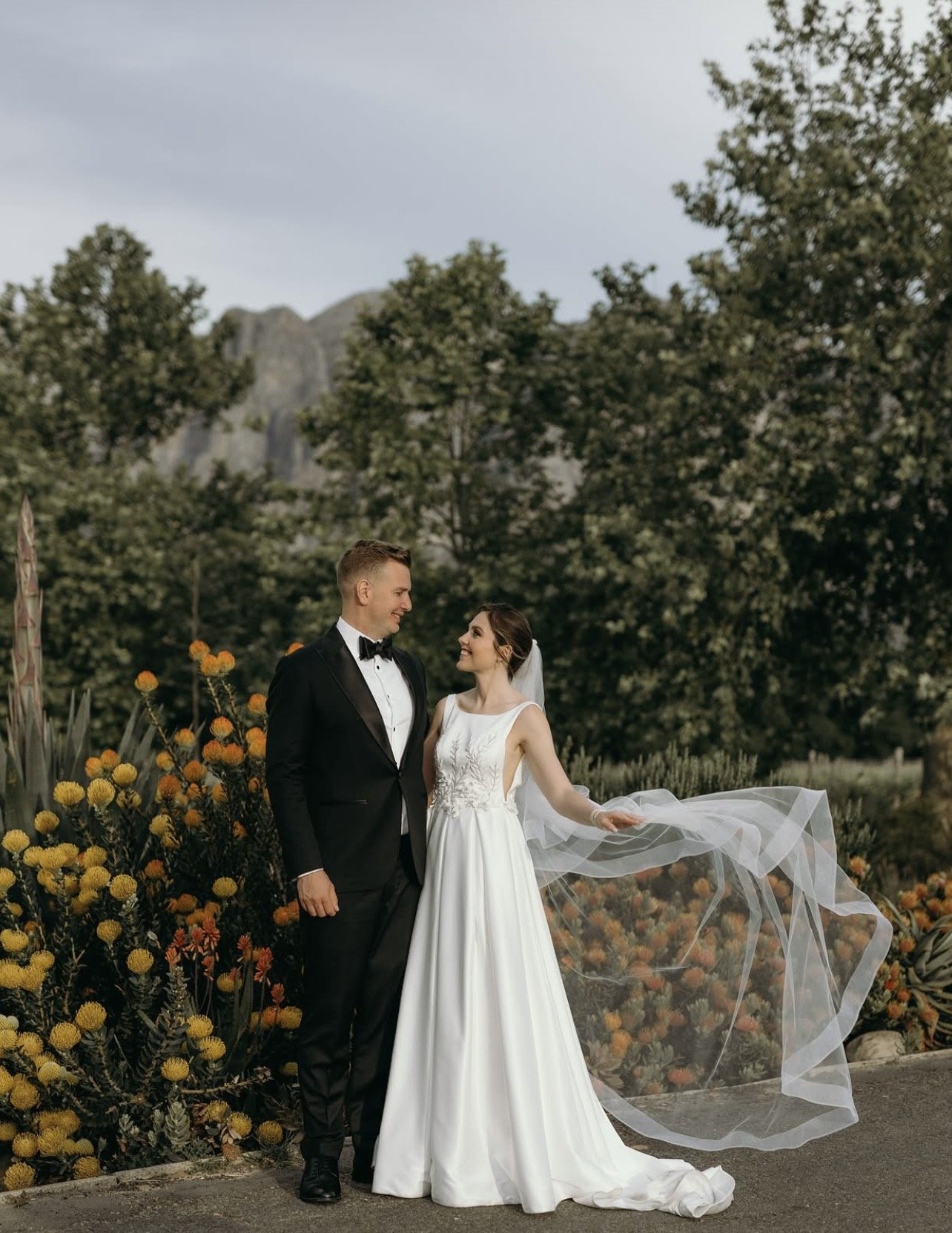 A bride and groom in wedding attire standing outdoors among yellow flowers, with trees and mountains in the background, smiling at each other.