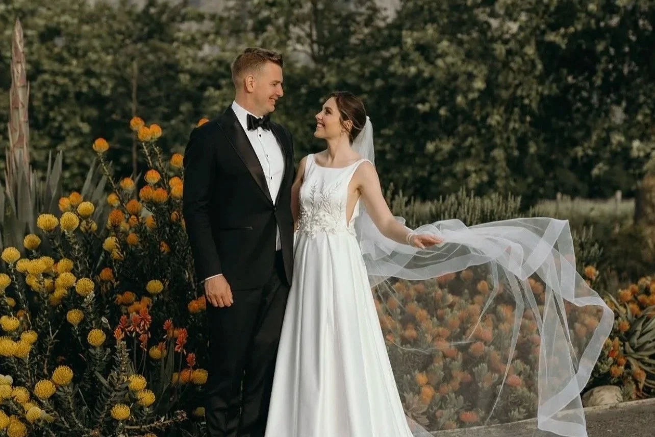 Bride and groom standing together outdoors during their wedding, with the bride holding her flowing veil and wearing a white dress, and the groom in a black tuxedo, surrounded by vibrant yellow and orange flowers.