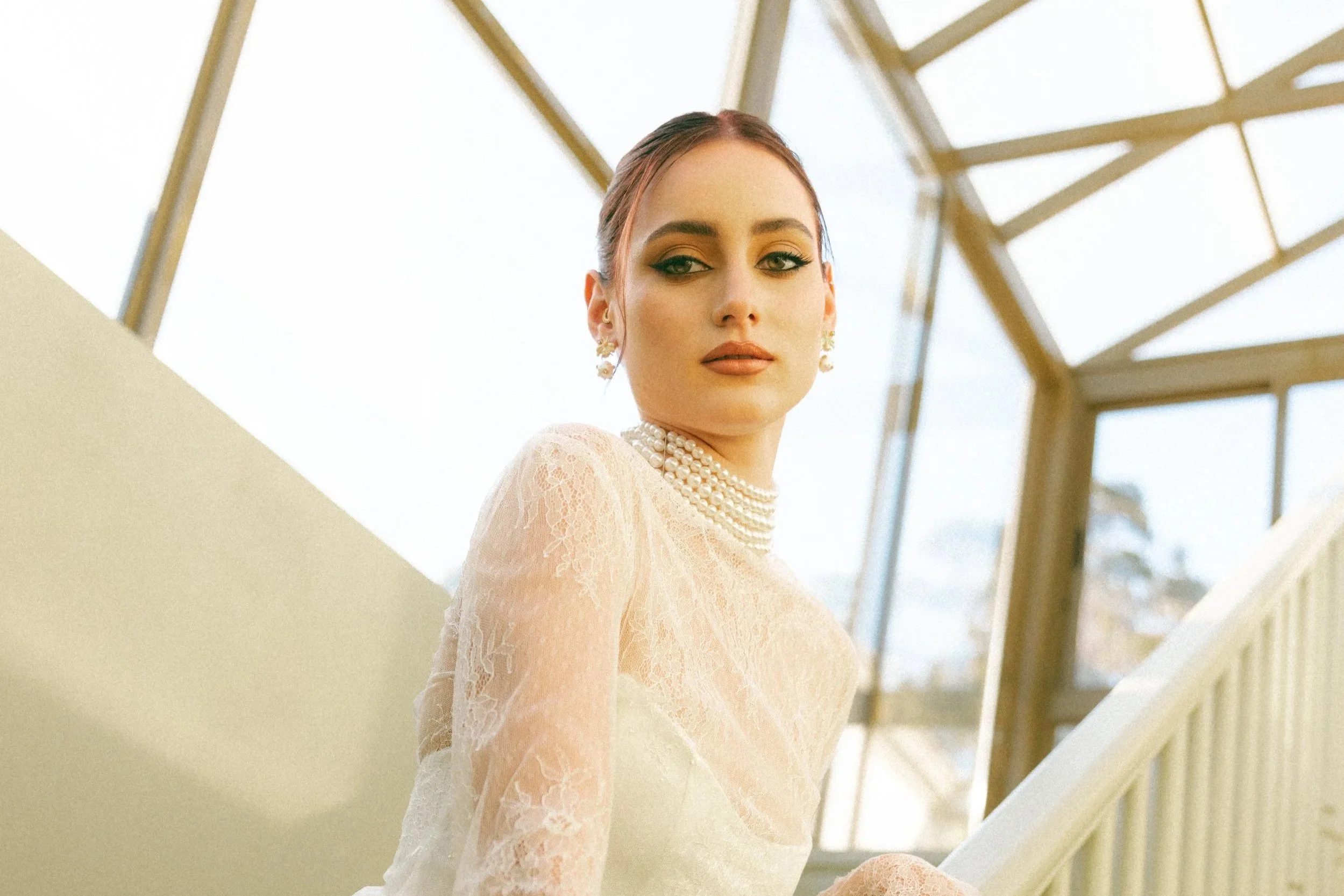 A woman in a lace dress and pearl jewelry standing inside a glass-structured building.