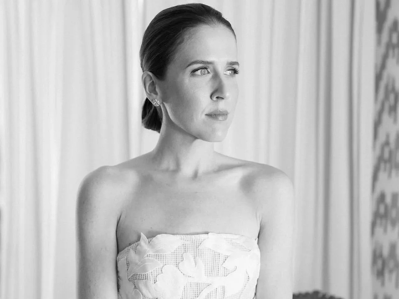 Black and white photo of a woman with short hair, wearing earrings and a strapless dress, looking to her right, in front of a curtain with a patterned curtain in the background.