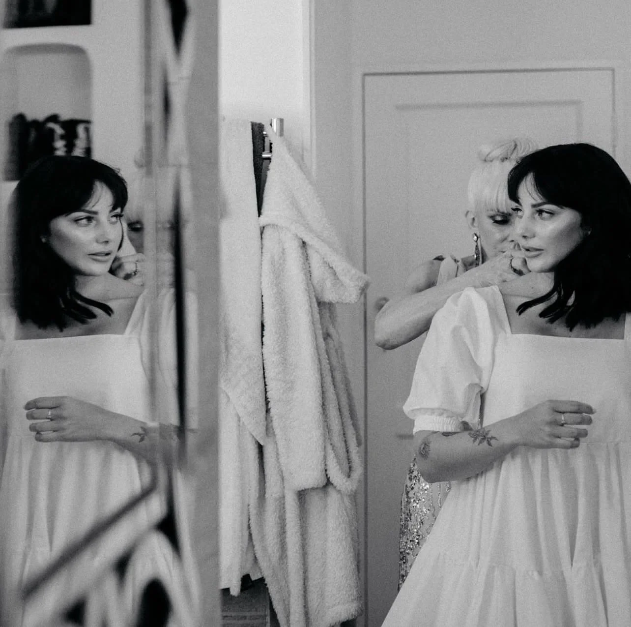 A woman in a white dress with dark hair looking at her reflection in a mirror while another woman adjusts her dress behind her in a room with towels hanging on the wall.