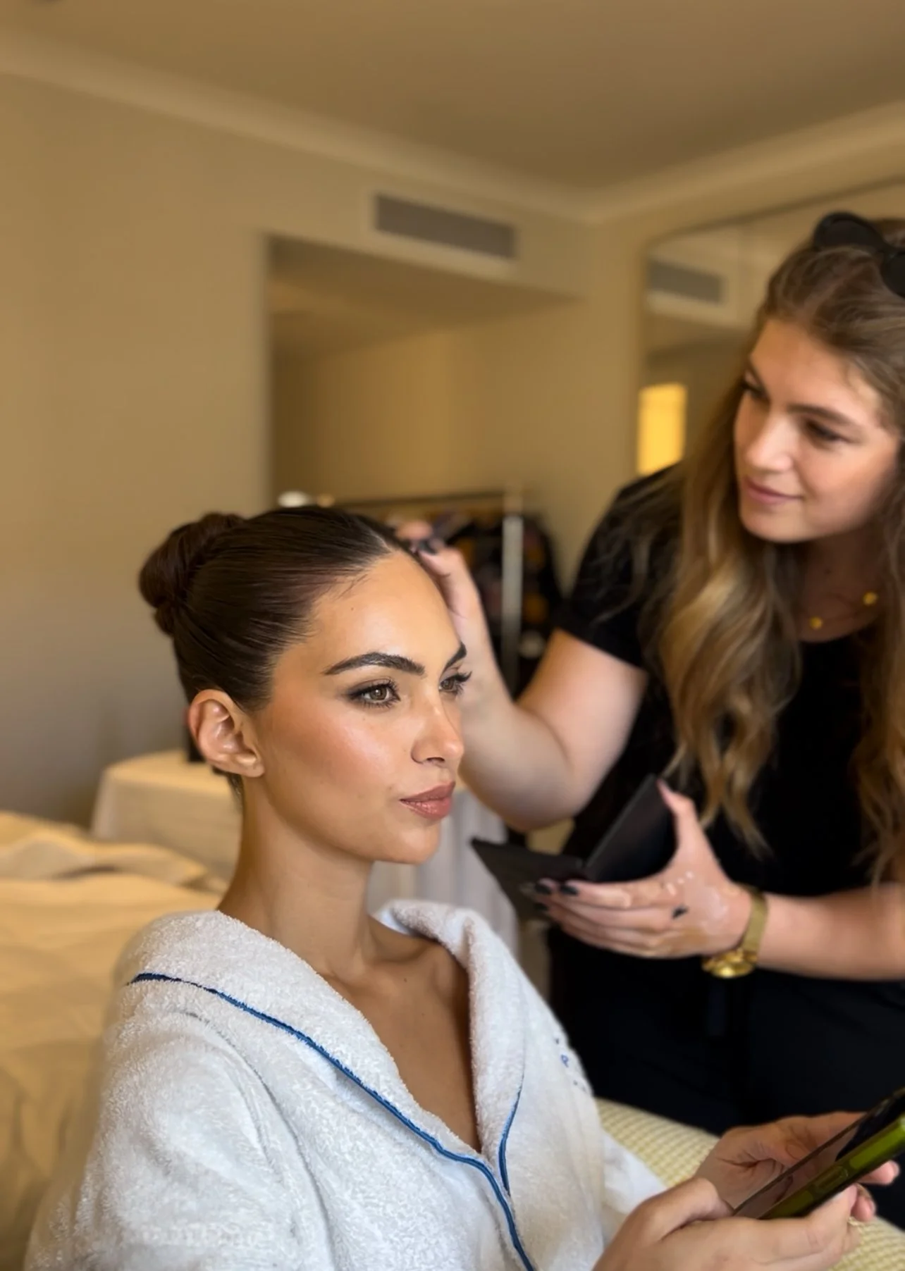 A woman in a white robe looks at her phone as another woman applies makeup or skincare nearby, with a blurred background of a bedroom or hotel room.