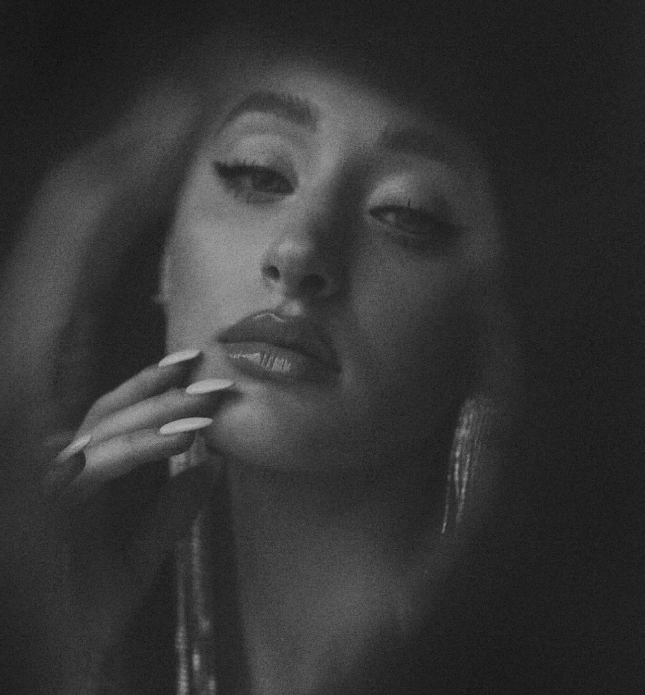 Black and white close-up portrait of a woman with long hair, makeup, and jewelry, looking at the camera with her hand resting near her chin.