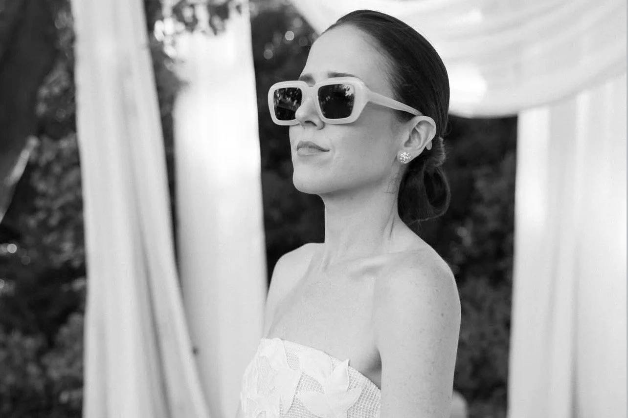 A woman wearing large sunglasses and earrings, dressed in a strapless top, standing outdoors near curtains or drapes.