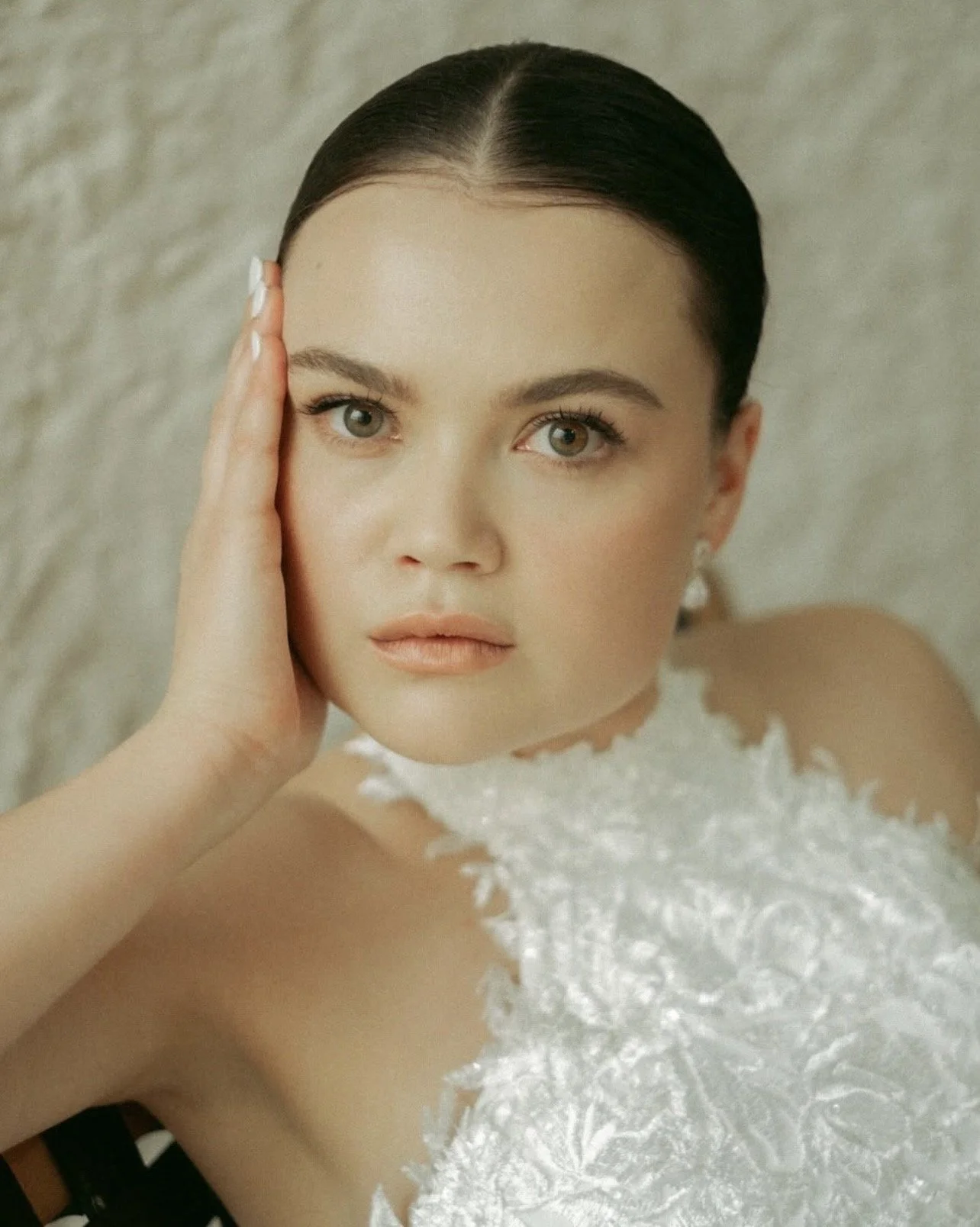 A woman with dark hair, styled back, resting her face on her left hand, looking directly at the camera, with a neutral expression. She is wearing a white, textured top or dress with floral details.