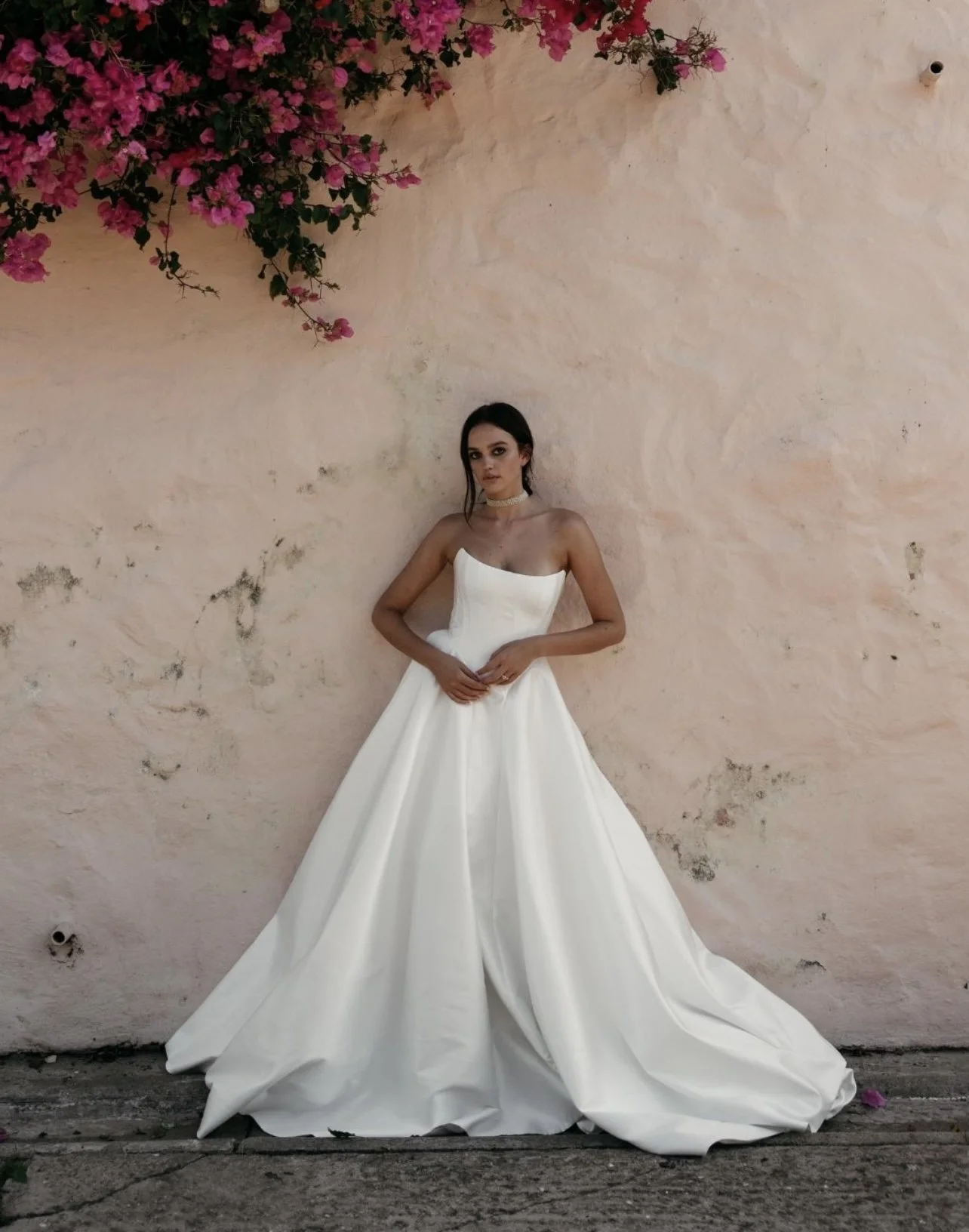 A woman in a white strapless wedding dress stands against a light pink wall with pink flowers hanging above her.