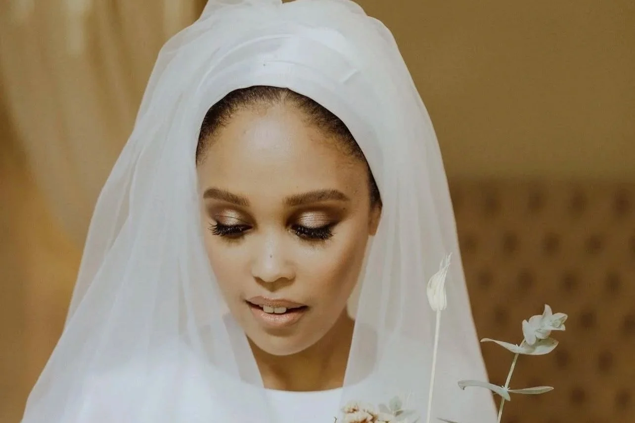 A woman wearing a white veil with makeup, holding a flower, and looking down.