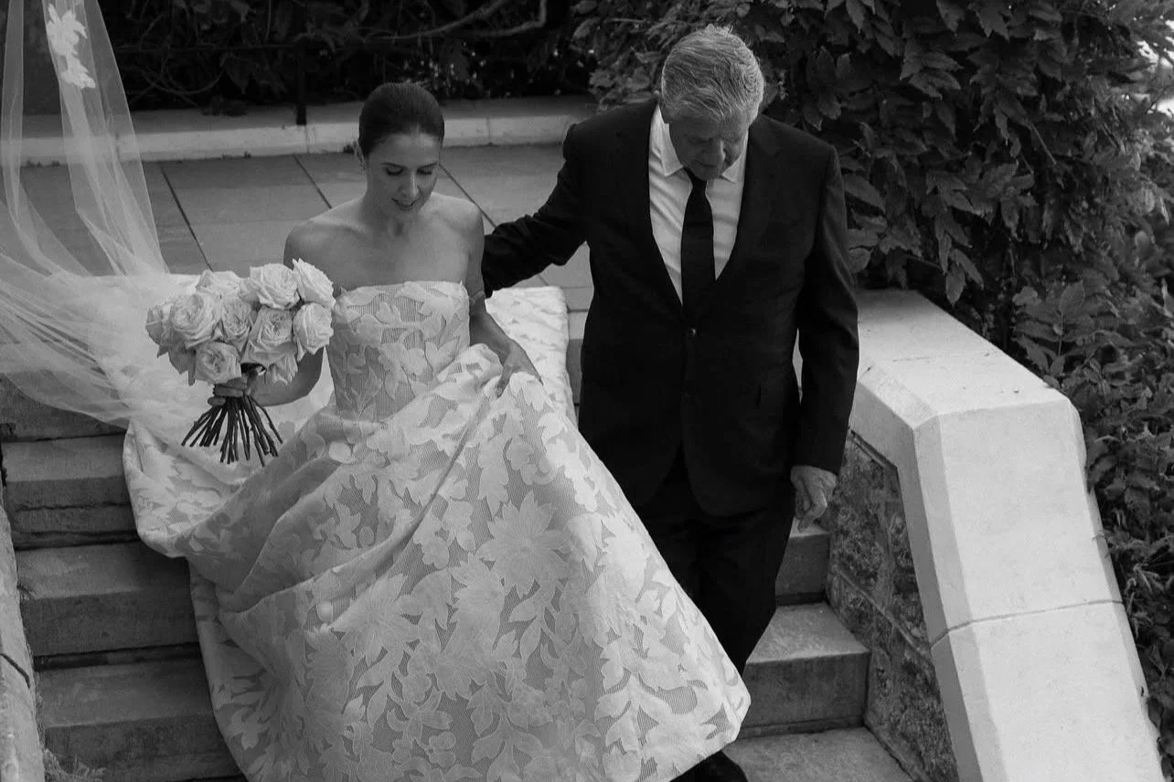 Black and white photo of a woman in a wedding dress holding a bouquet of roses being assisted down stairs by a man in a suit.