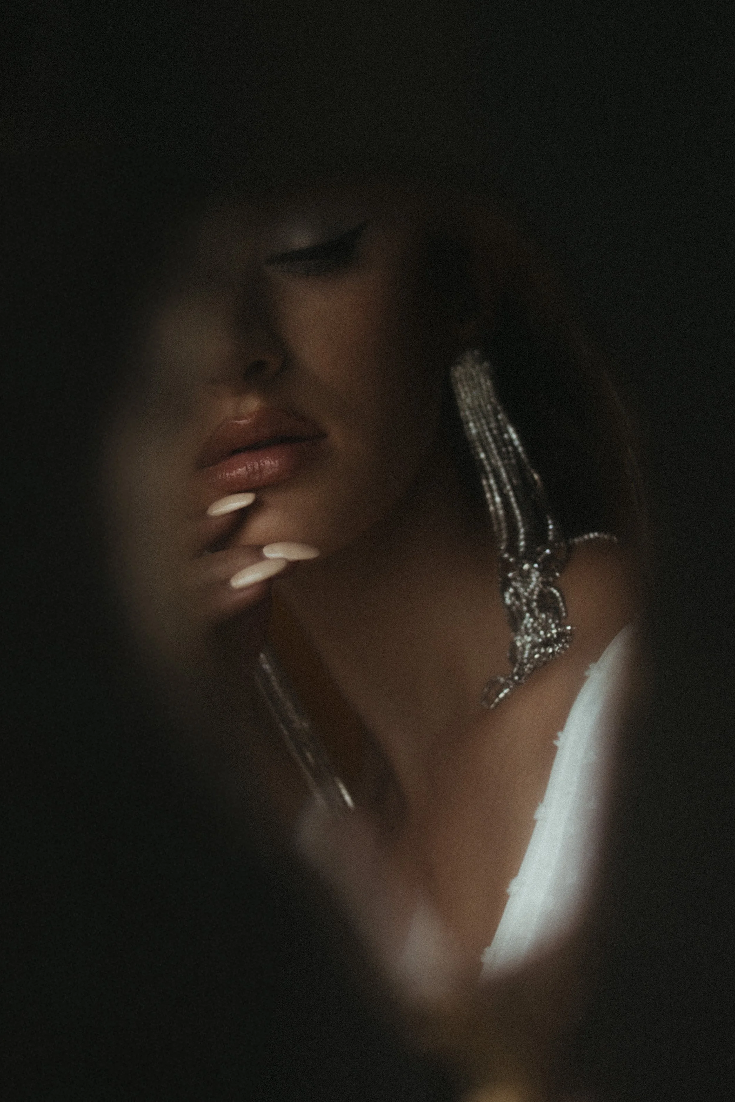 A woman with makeup, wearing long, sparkly earrings and a white garment, touching her chin with her fingers, looking down, partly obscured by darkness or shadow.