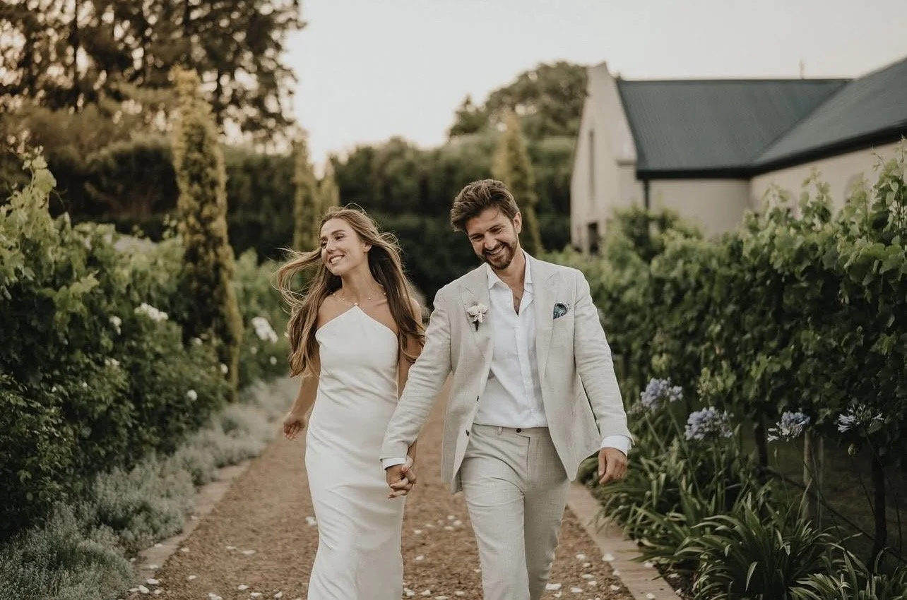 A newlywed couple walking hand in hand through a garden path, smiling and enjoying their day. Makeup by Andrea van Den Houten France based makeup artist 