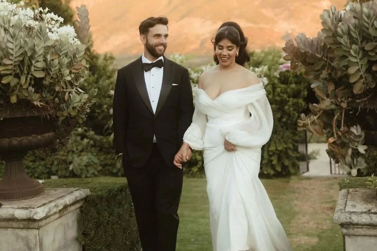 A couple in wedding attire walking outdoors, with the man in a black tuxedo and the woman in a white off-shoulder wedding gown, surrounded by greenery and large floral arrangements. Make Up by Paris based Makeup Artist Andrea van Den Houten