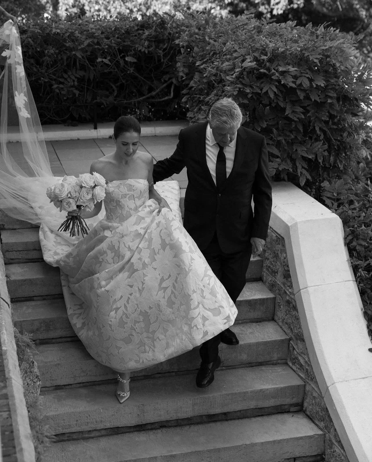 A bride in a floral wedding gown and heels sitting on outdoor stairs holding a bouquet of roses, being assisted by a man in a dark suit descending the stairs.