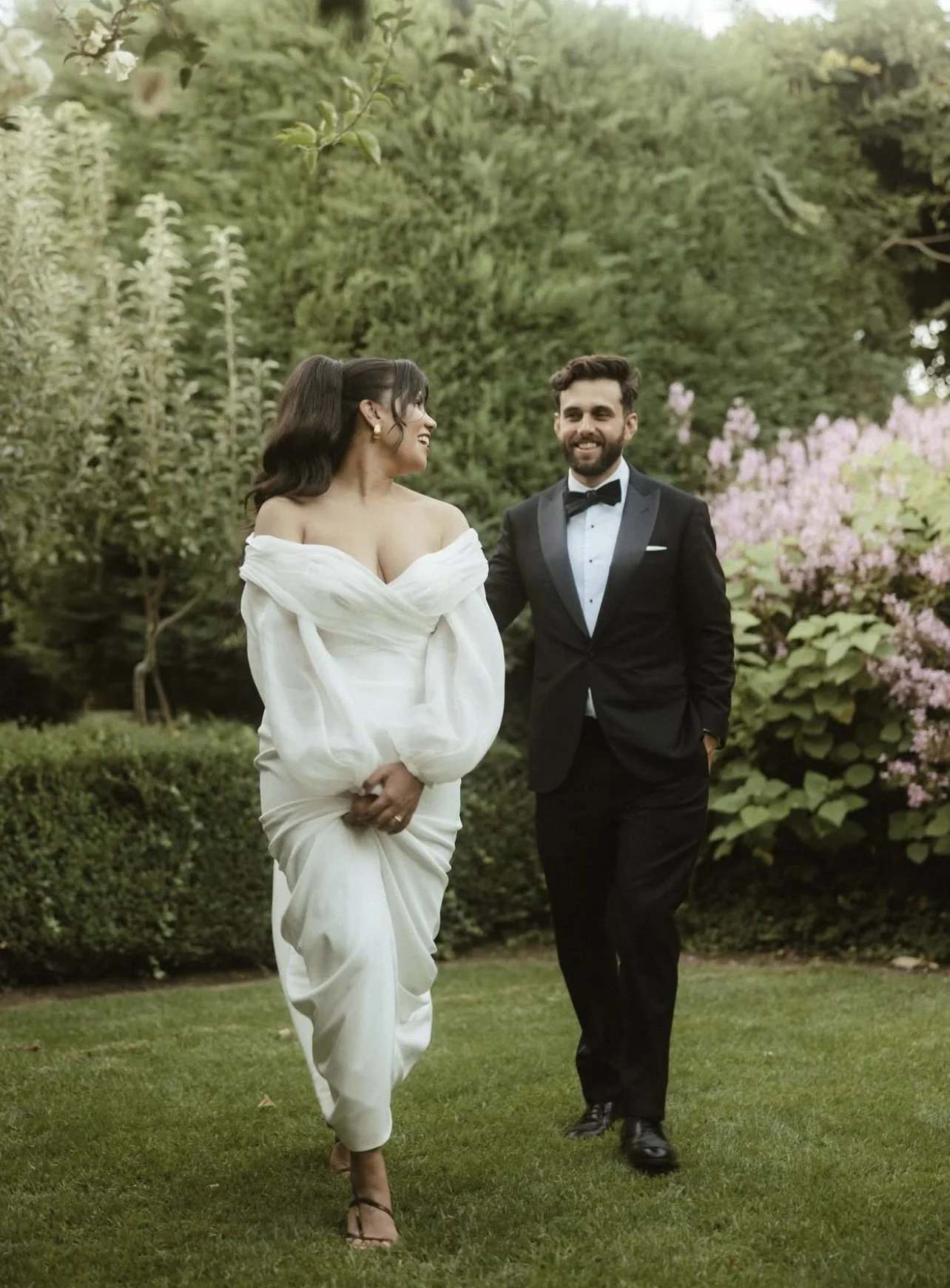 A couple in wedding attire walking outdoors on a grass lawn with greenery and flowering bushes in the background. The woman is wearing a white off-shoulder wedding dress, and the man is in a black tuxedo with a bow tie.