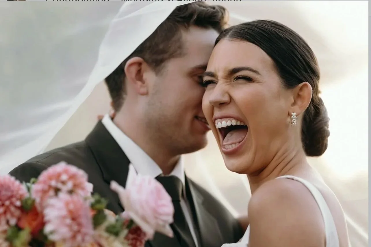 Joyful bride and groom laughing closely on their wedding day, with flowers in the foreground.