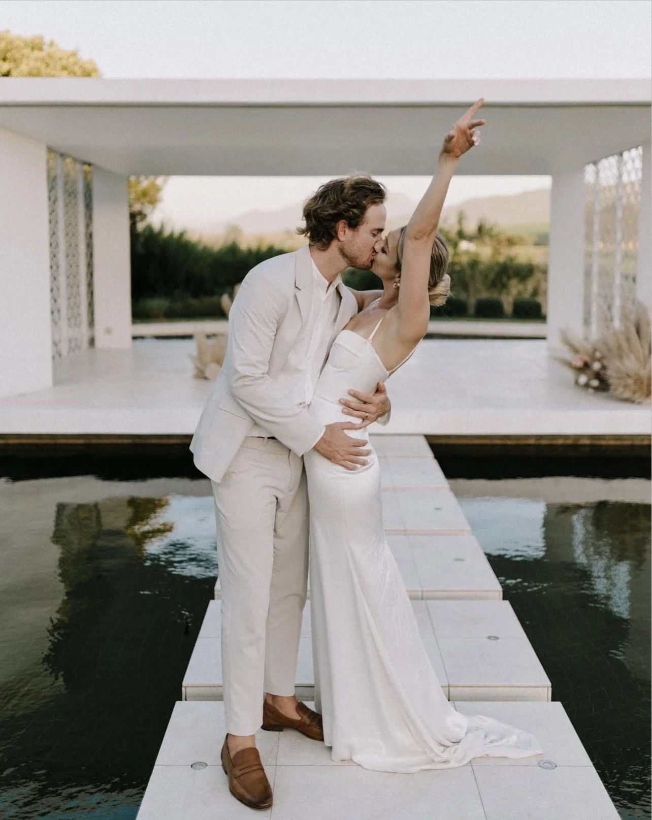 A newlywed couple sharing a kiss on a modern, minimalist outdoor platform over water, with one arm raised in celebration and lush greenery in the background.