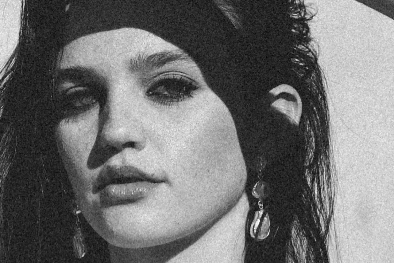 Close-up of a woman with dark hair, wearing earrings, looking into the camera in black and white.