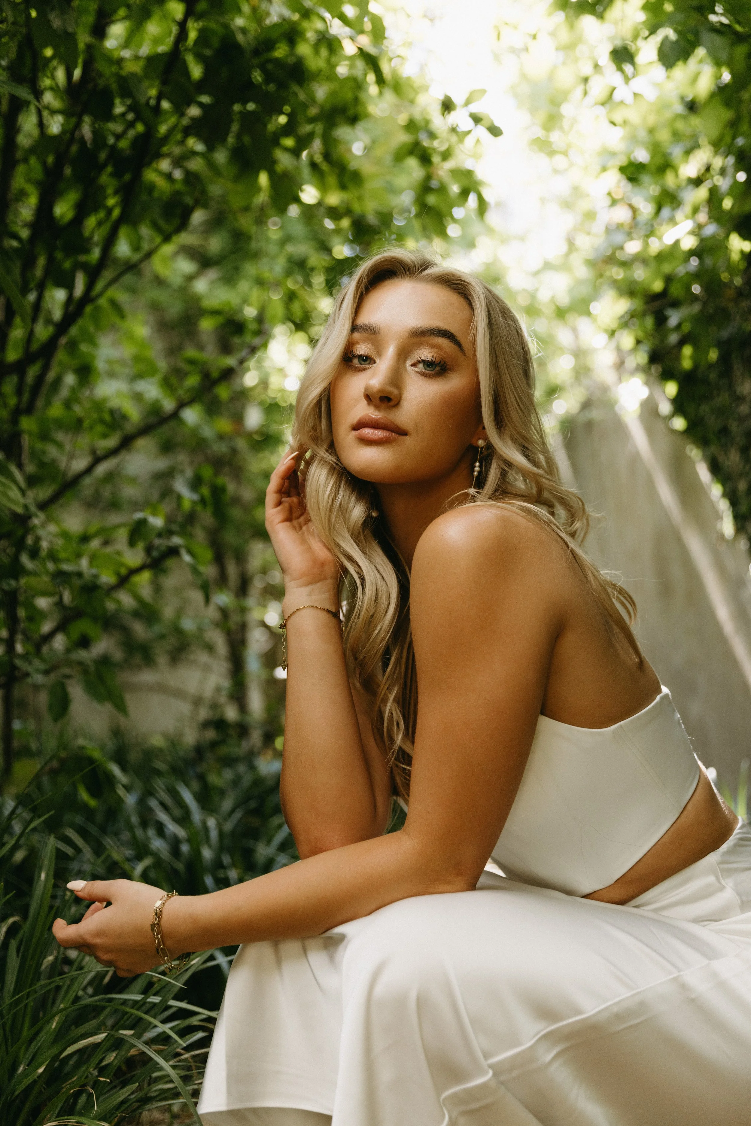 A young woman with long blonde hair wearing a white strapless top and matching white skirt, sitting outdoors among lush greenery, looking at the camera.