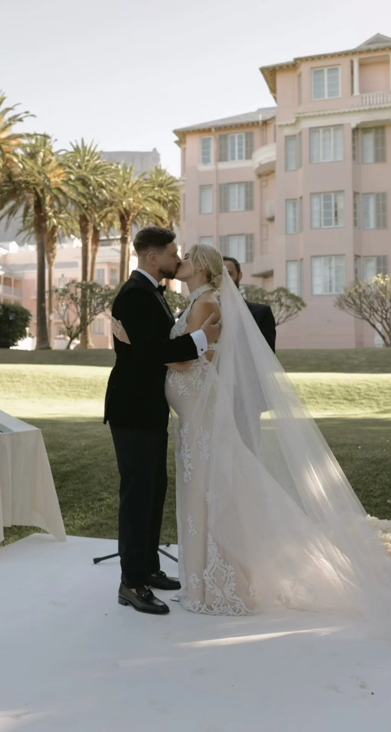 A bride and groom kiss during their wedding ceremony outdoors, with a pastel pink building and palm trees in the background.