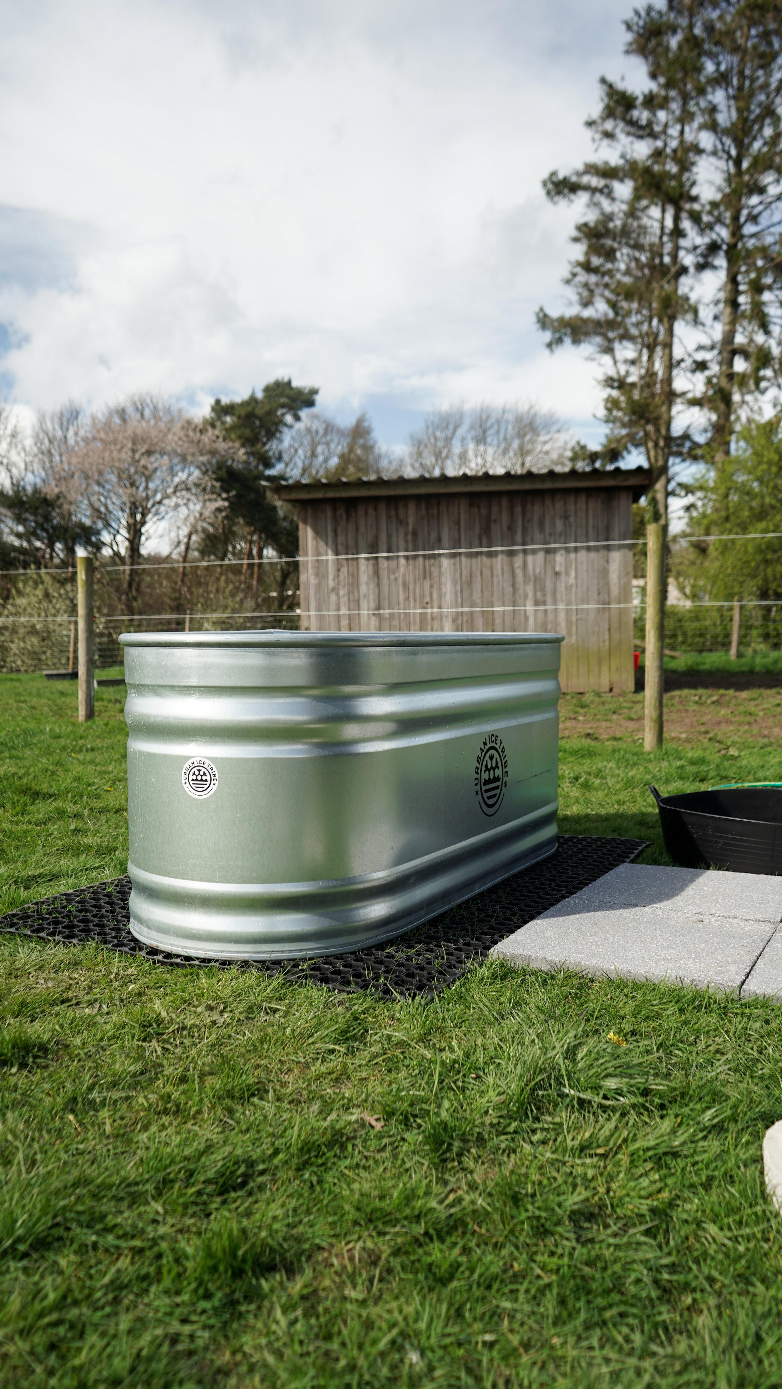 A metal planter on a patch of grass with a black mat underneath. In the background are trees, a wooden shed, and a cloudy sky.
