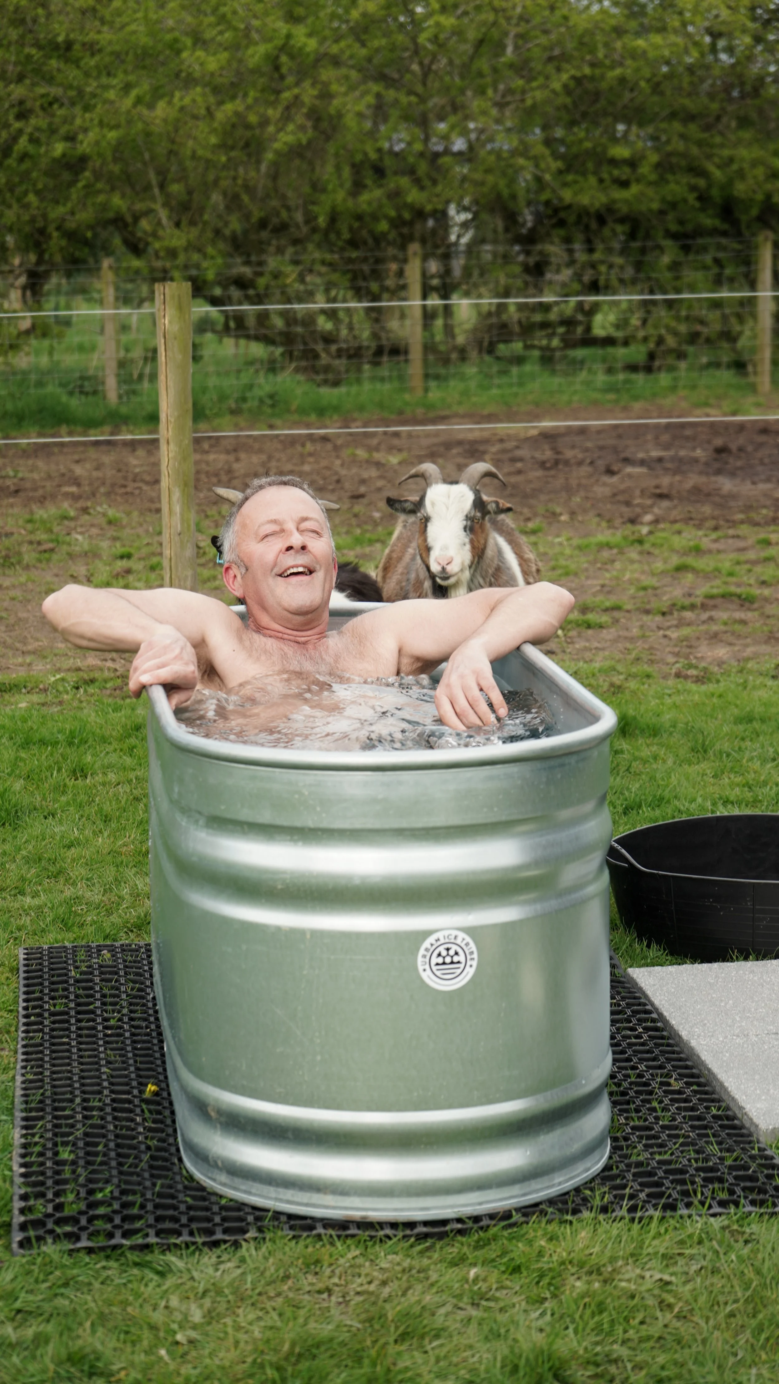 A man relaxing in a metal tub filled with water outdoors, with a goat standing behind him, in a grassy area with a fence and trees in the background.