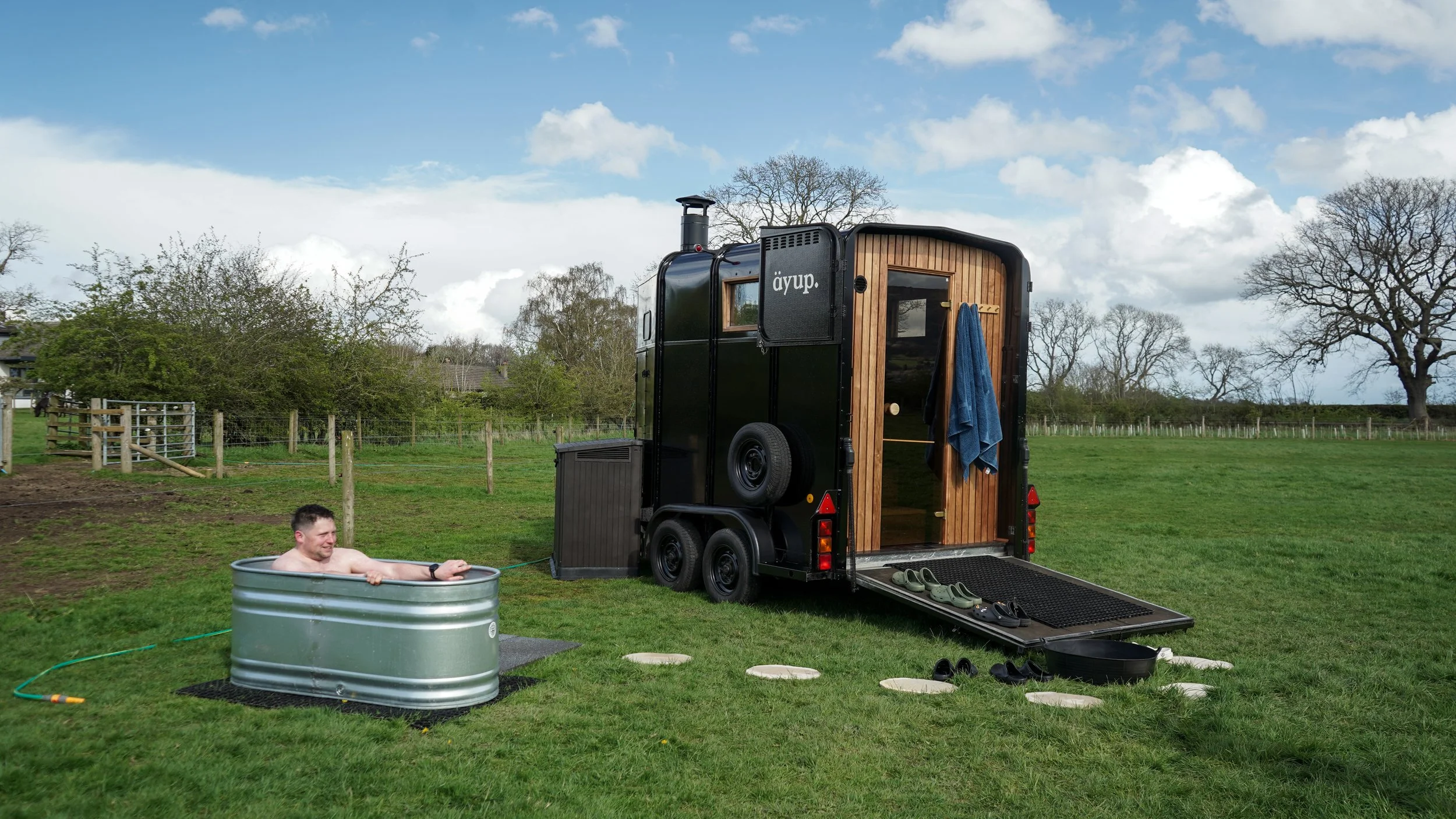 A man relaxing in a small metal hot tub outdoors on green grass, connected to a black tiny house on wheels with a wooden door and a window, set in a rural area with trees and a blue sky with clouds.