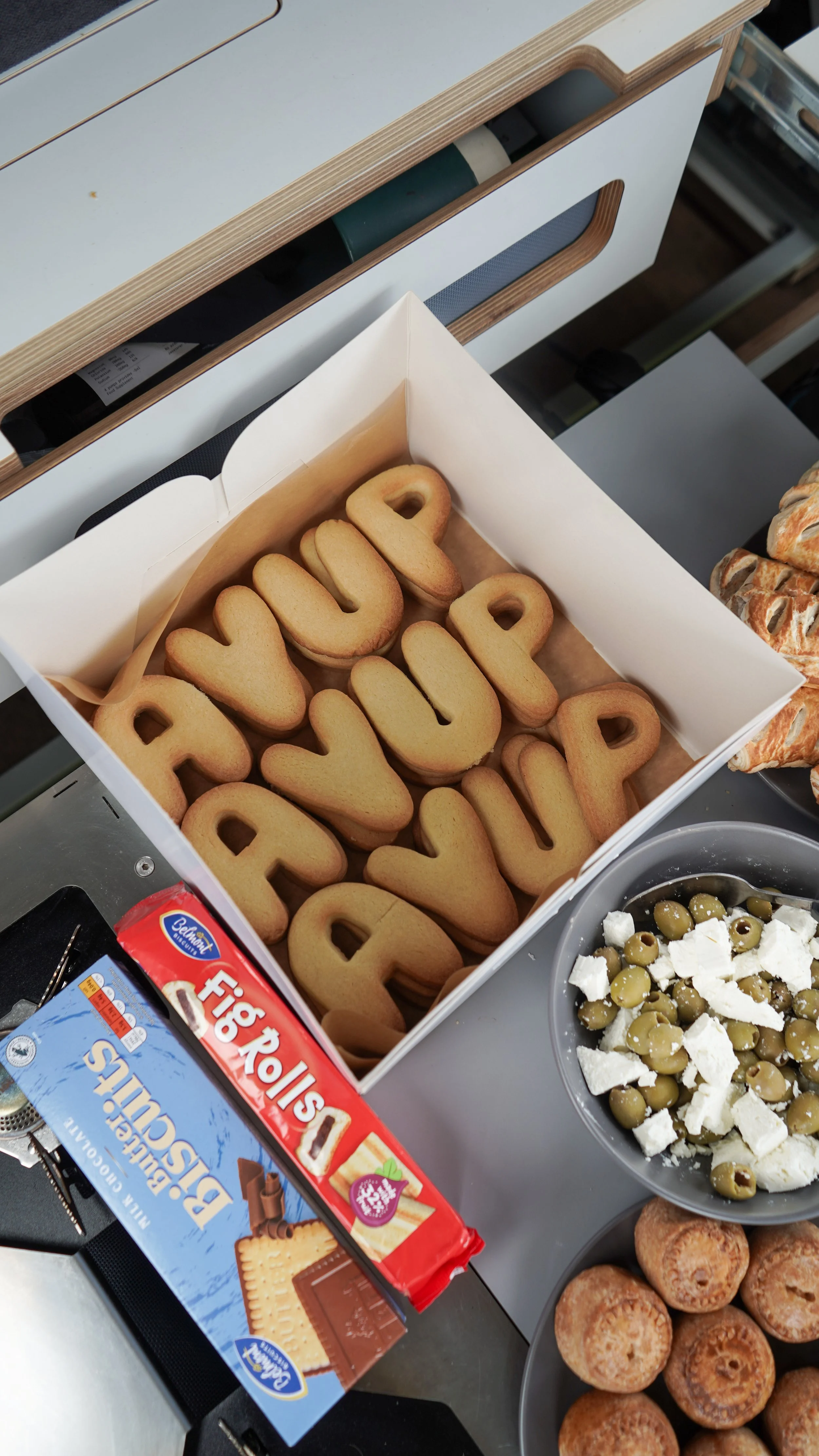 A box of cookies spelling out 'HAPPY BIRTHDAY' with a tray of Greek salad and a plate of pastries on a table.