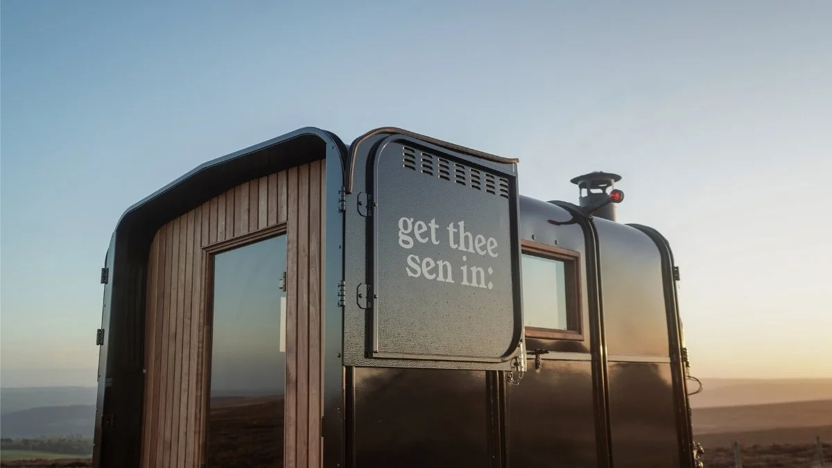 A black trailer with wooden accents and a small window, outdoors during sunset, with a weathered sign that reads "get thee sen in:"