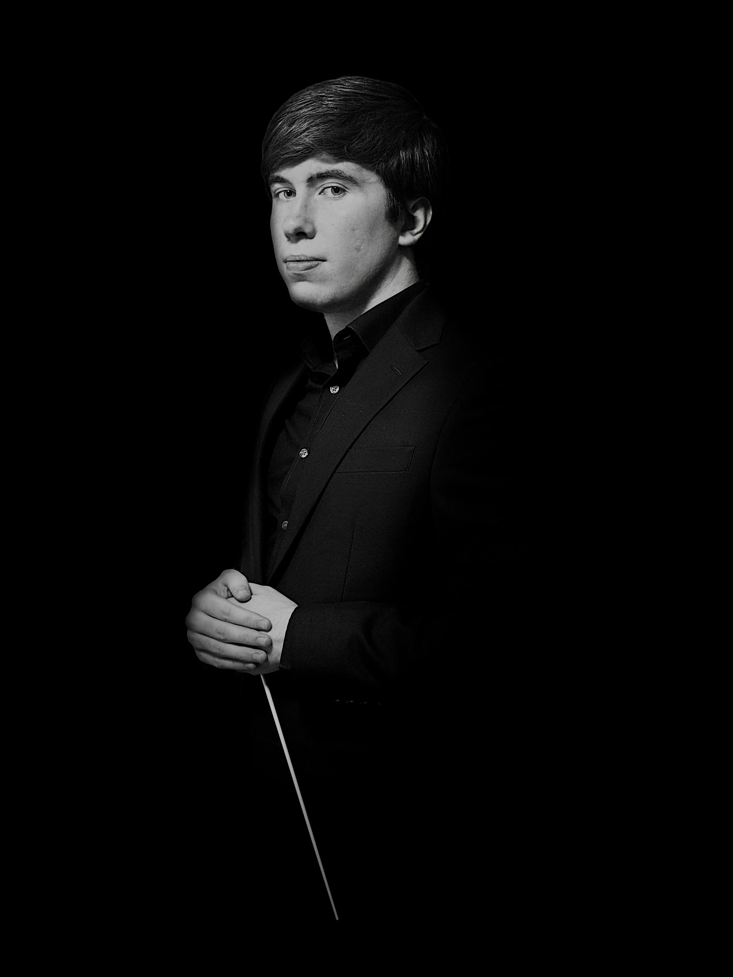Black-and-white portrait of a young man in a suit, holding a conductor's baton, against a dark background.