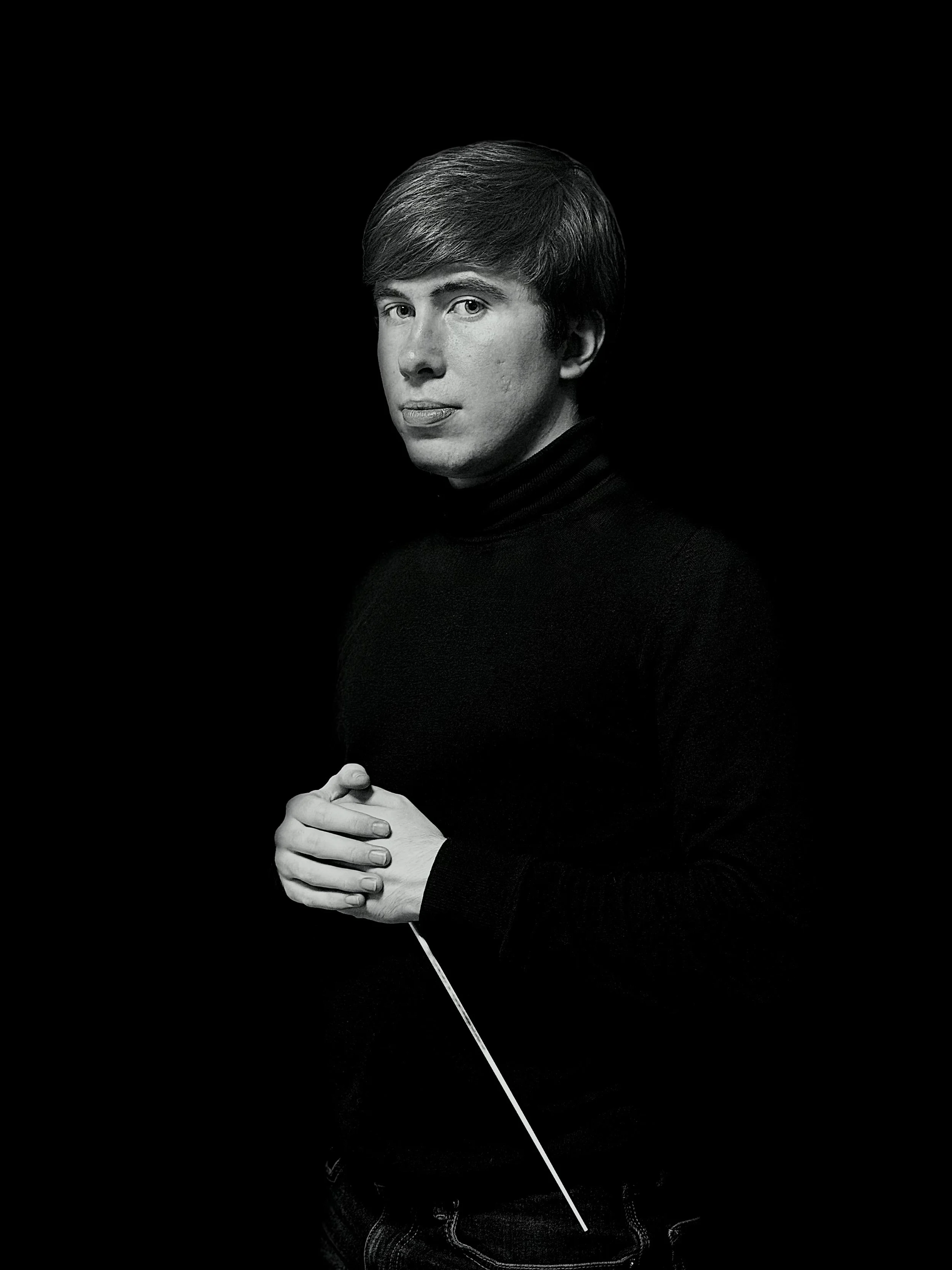 Black-and-white portrait of a young man in a black turtleneck, holding a conductor's baton, against a dark background.