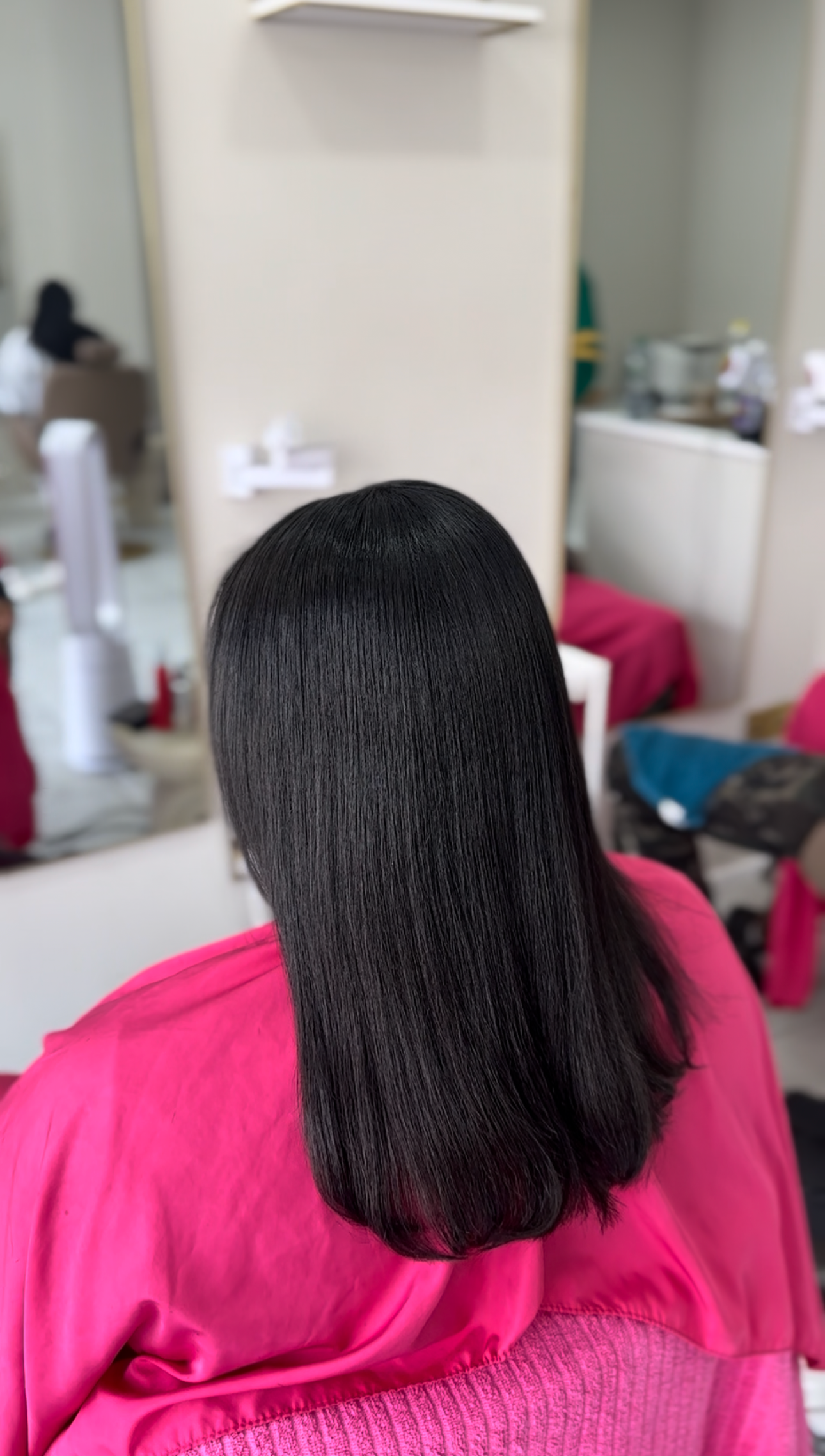 A woman with long, straight, black hair sitting in a salon chair wearing a pink cape, with a mirror and hairdressing tools visible in the background.