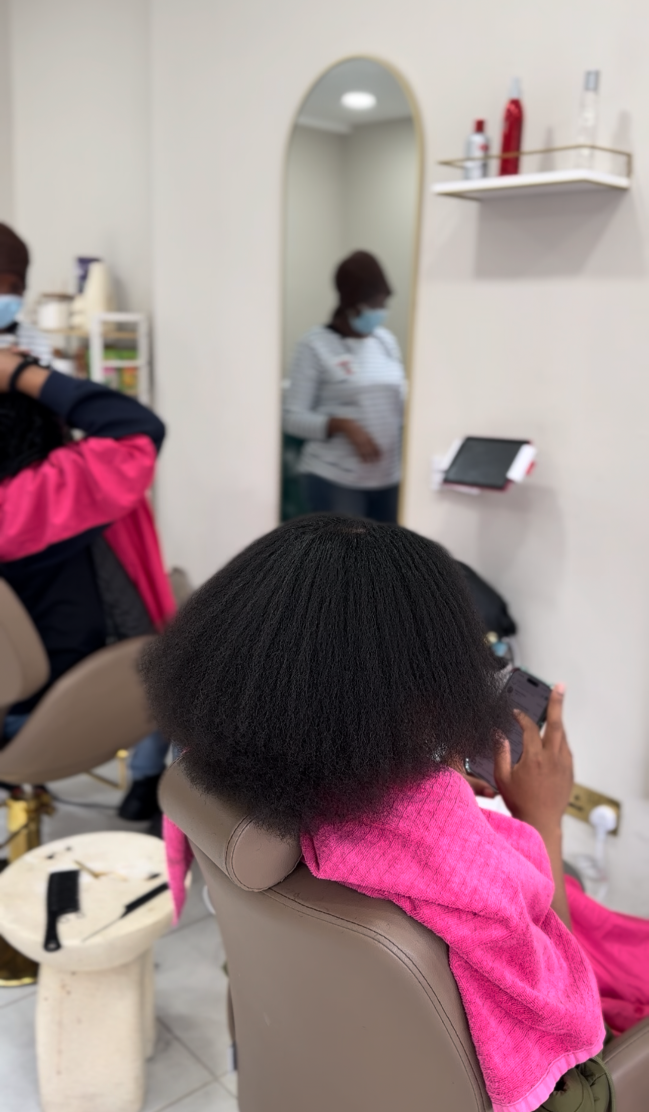 A woman sitting in a salon chair with a pink towel around her neck, using her phone. A mirror and shelves with hair products are visible in the background.