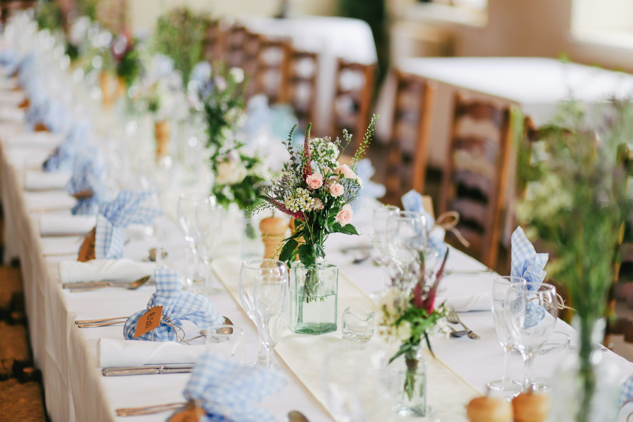 A long dining table set for a celebration with floral centerpieces, blue checkered napkins, and glassware in a well-lit room.