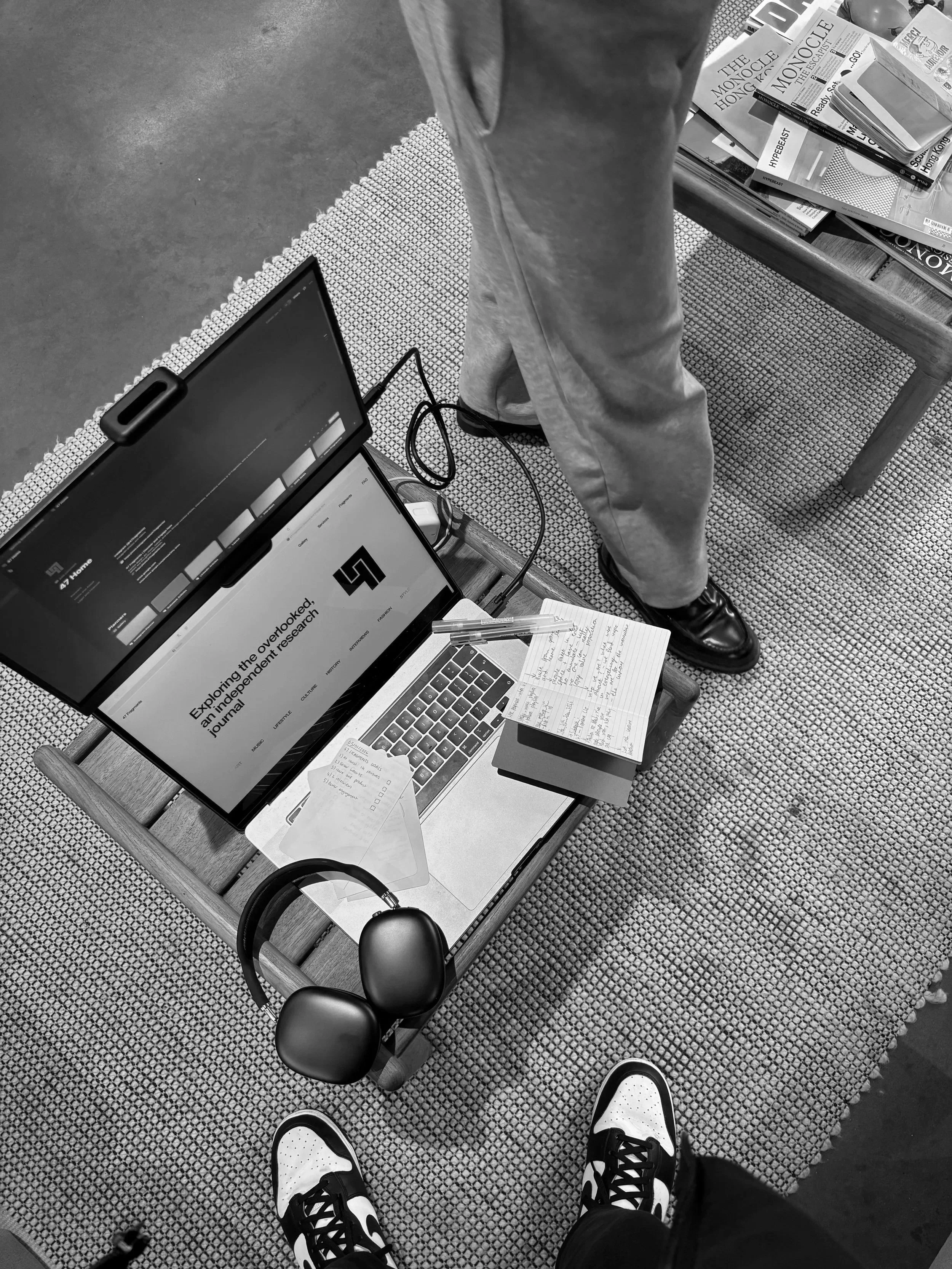 A top-down view of a workspace with a person's legs, a laptop, a tablet, some notebooks with handwritten notes, a pair of headphones, and a book on a wooden table. Additional books are on a second table nearby.