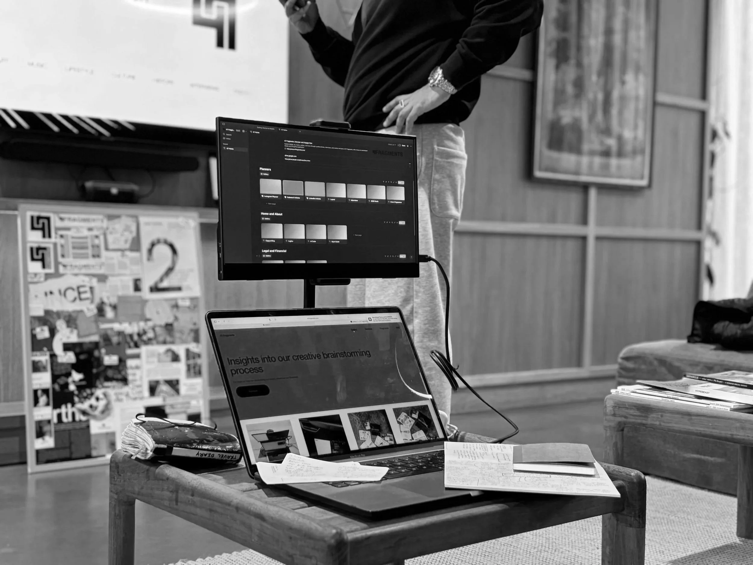 A workspace with a wooden table holding a laptop, a notebook, and a piece of paper. A monitor is mounted above, displaying a dark-themed interface with color swatches. A person stands in the background, wearing a black hoodie and gray sweatpants, par