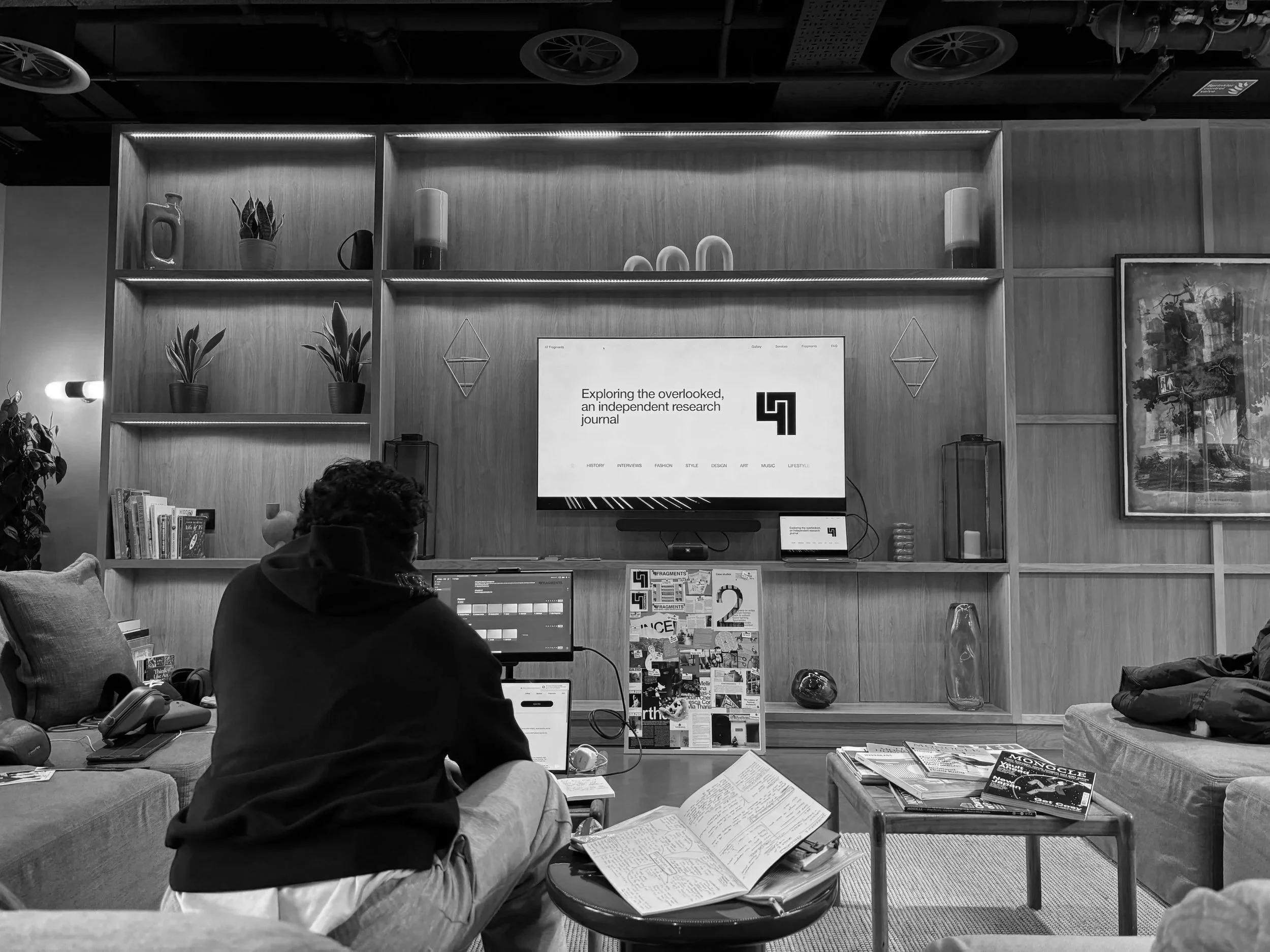 A person sitting in a cozy living room or lounge, working on a laptop with books and magazines on a coffee table, facing a large wooden wall with a TV screen displaying a research journal website, surrounded by decorative plants and artwork.
