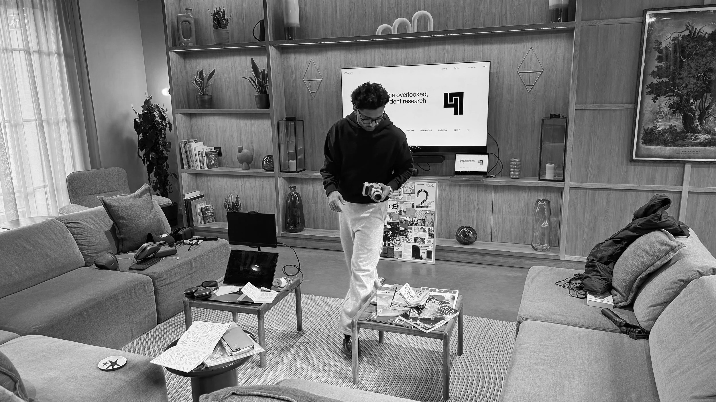 A man stands in a cozy living room holding a camera. The room features a large sectional sofa with pillows, a coffee table with magazines and notes, a television mounted on a wooden wall unit, and various decorative items. There are books, plants, an