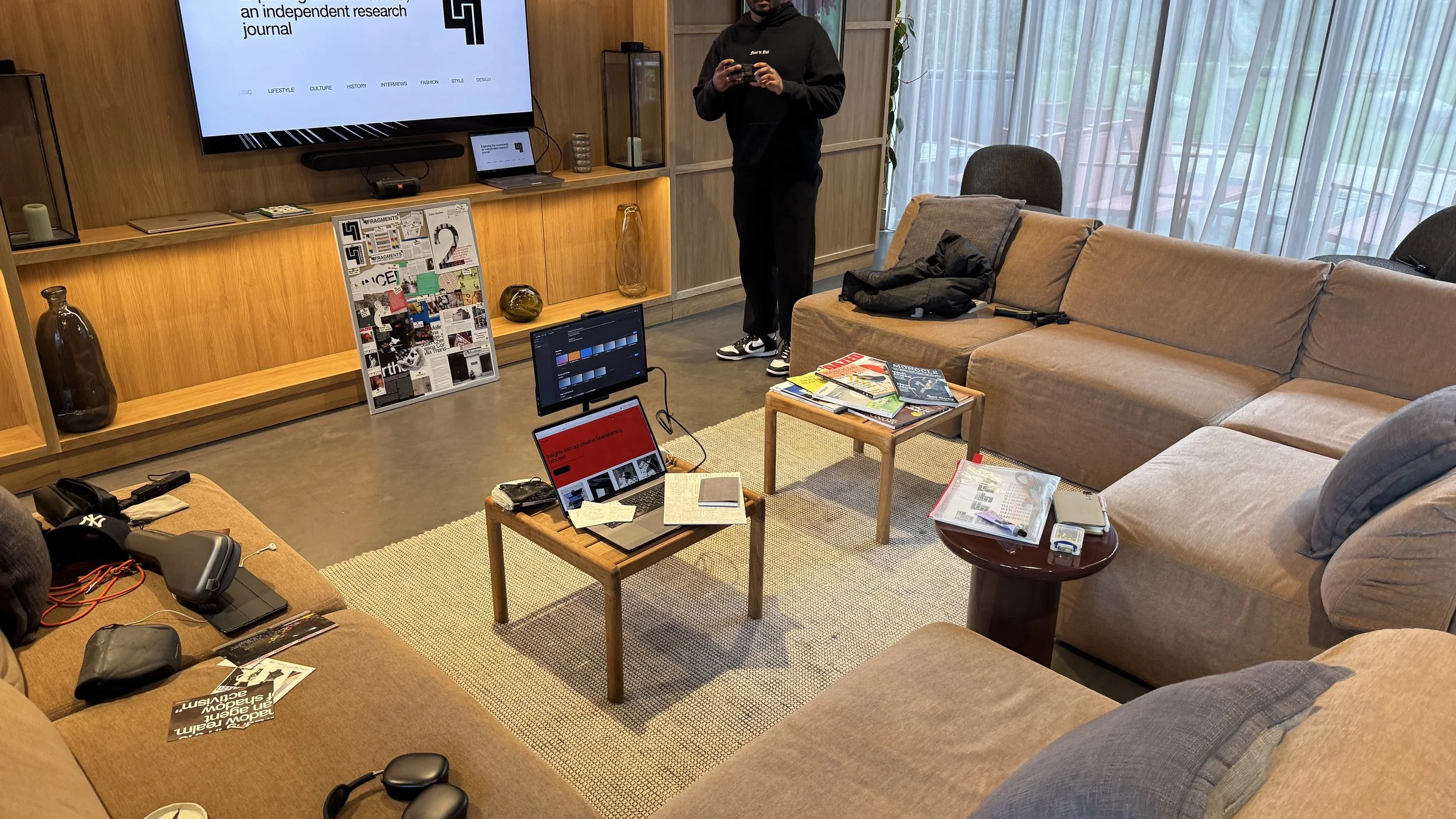 Living room with beige sectional sofa, coffee tables with magazines, laptops, and notebooks, a person standing holding a phone, large window with curtains in the background, TV mounted on a wooden wall, decorative vases, and various electronic devices and personal items on the furniture.