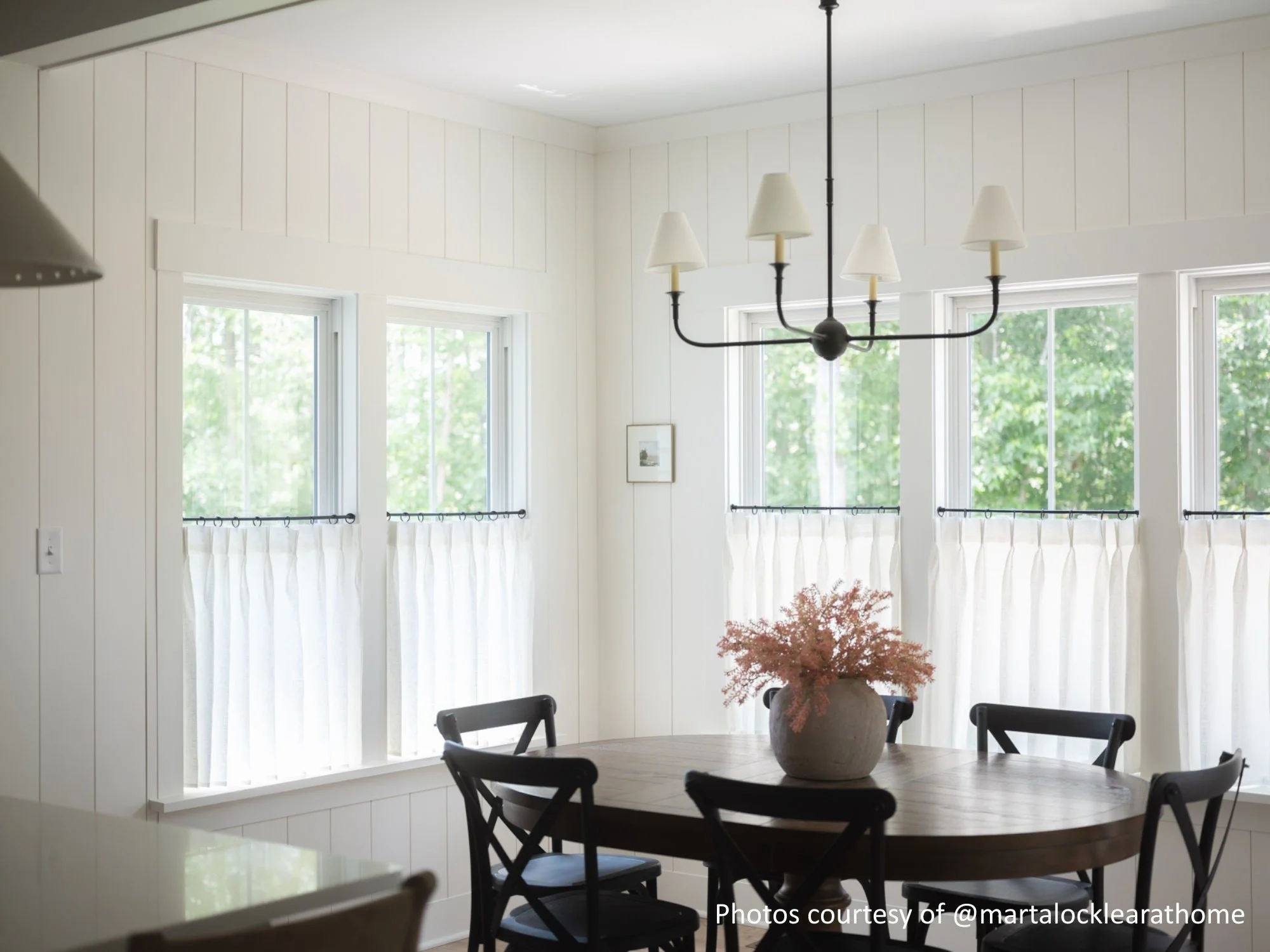 A bright dining area with white walls and large windows with white curtains, a black chandelier, a wooden round table with a pinkish flower arrangement, and black chairs.