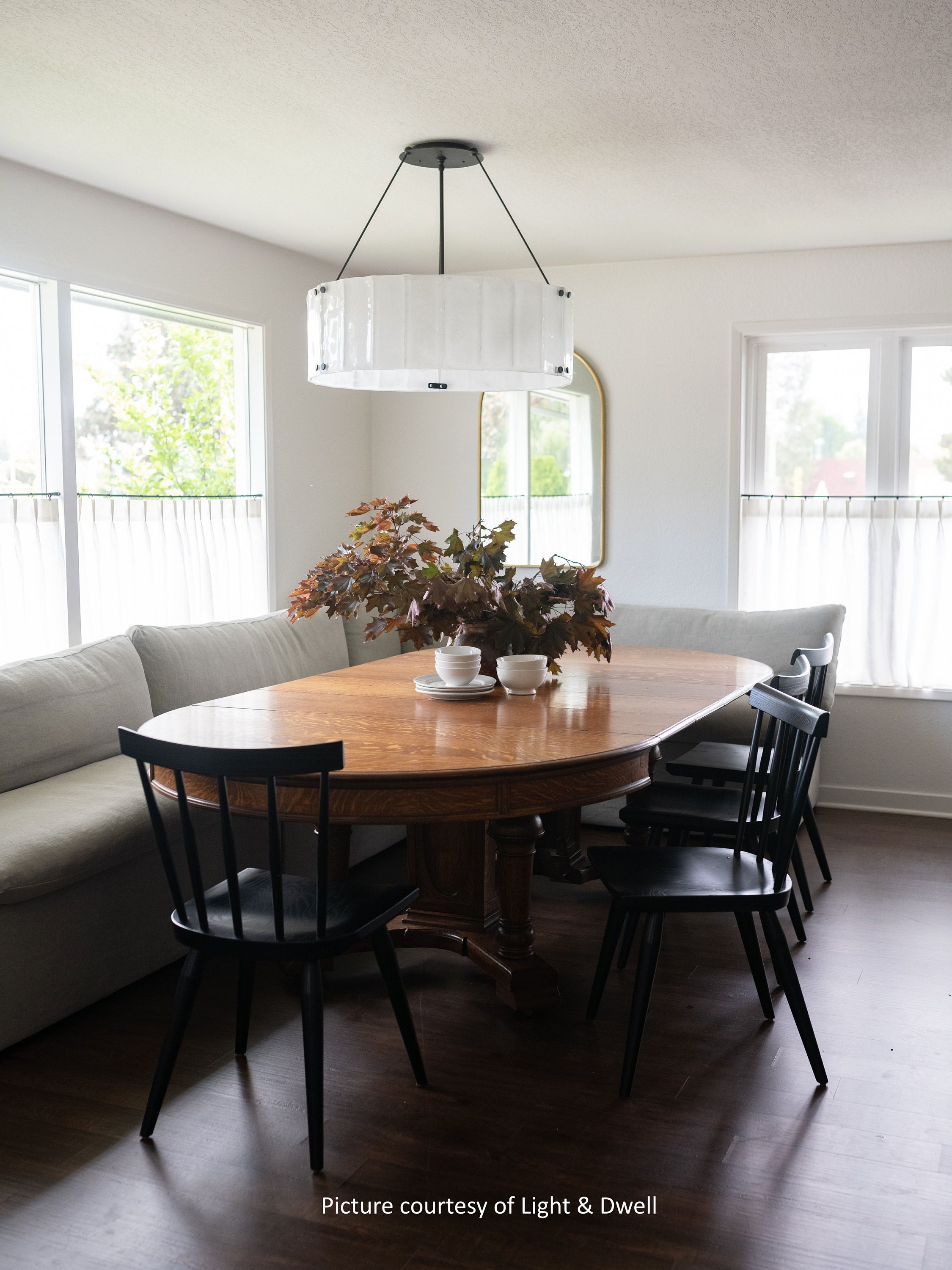 A dining room with a wooden oval table, a beige couch along the wall, black chairs around the table, a white pendant light hanging from the ceiling, and windows with white curtains. There are some houseplants on the table.