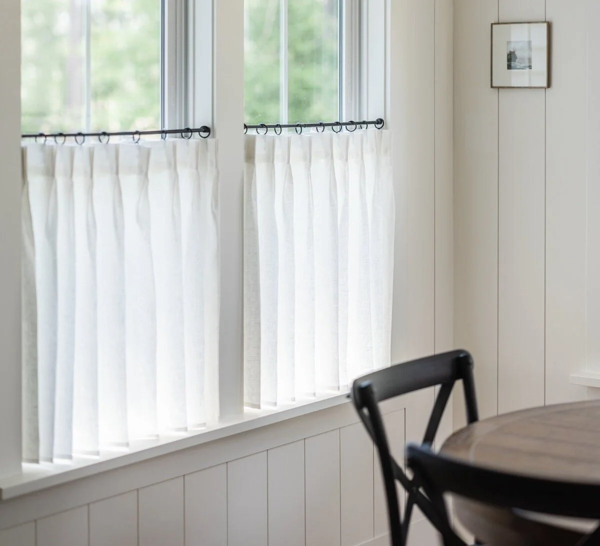 Bright kitchen window with white curtains and a black chair near a wooden table, with a framed picture on the wall.