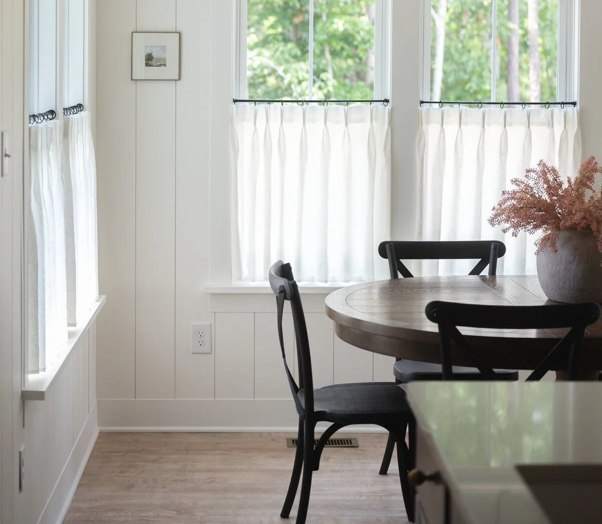 A cozy dining nook with a round wooden table, three black chairs, a large window with white curtains, a small picture on the wall, pink flowers in a vase, and natural light flooding the space.
