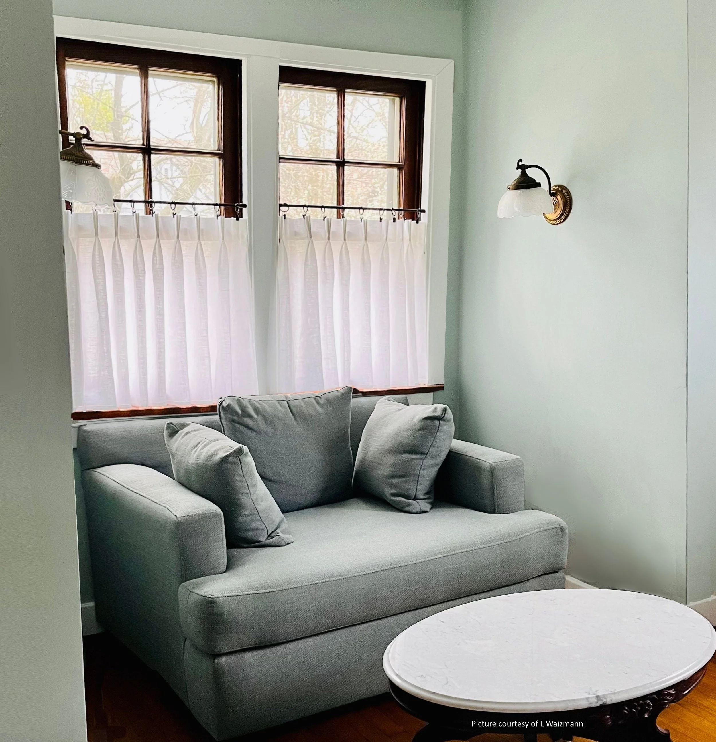 A cozy living room corner with a light gray sofa, three matching cushions, a white round marble-topped coffee table, a large window with white curtains, and wall-mounted vintage-style light fixtures.